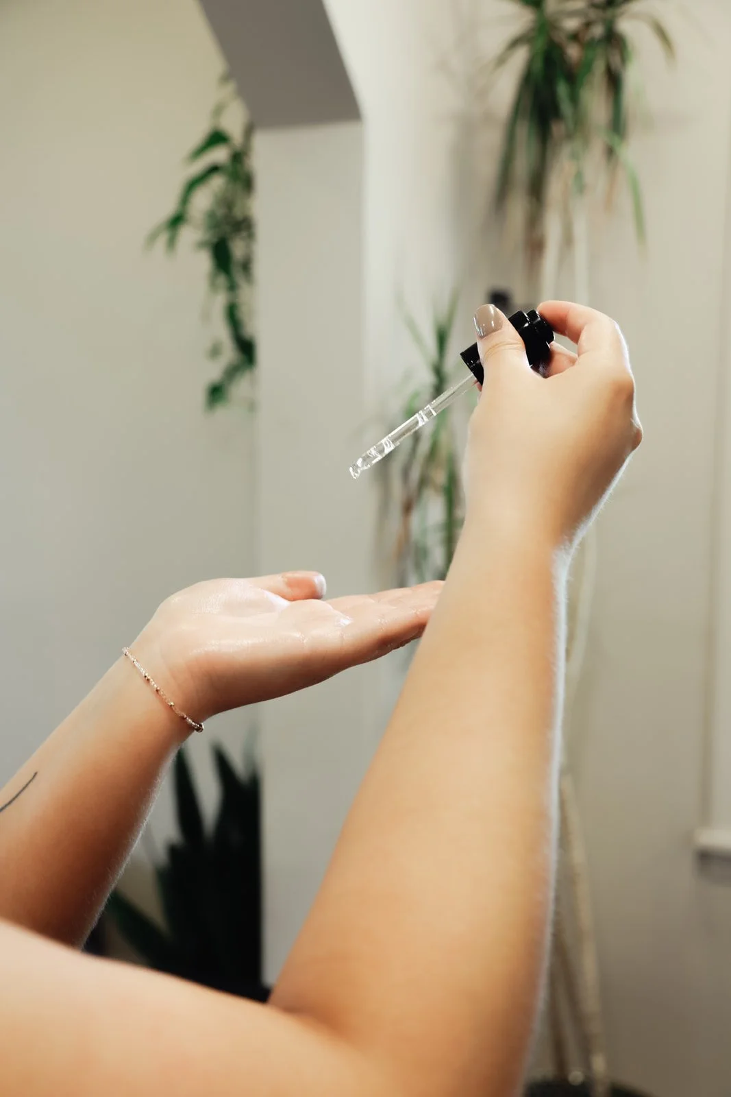 Person holding a dropper with liquid, dispensing it into palm of the other hand indoors with plants in the background.