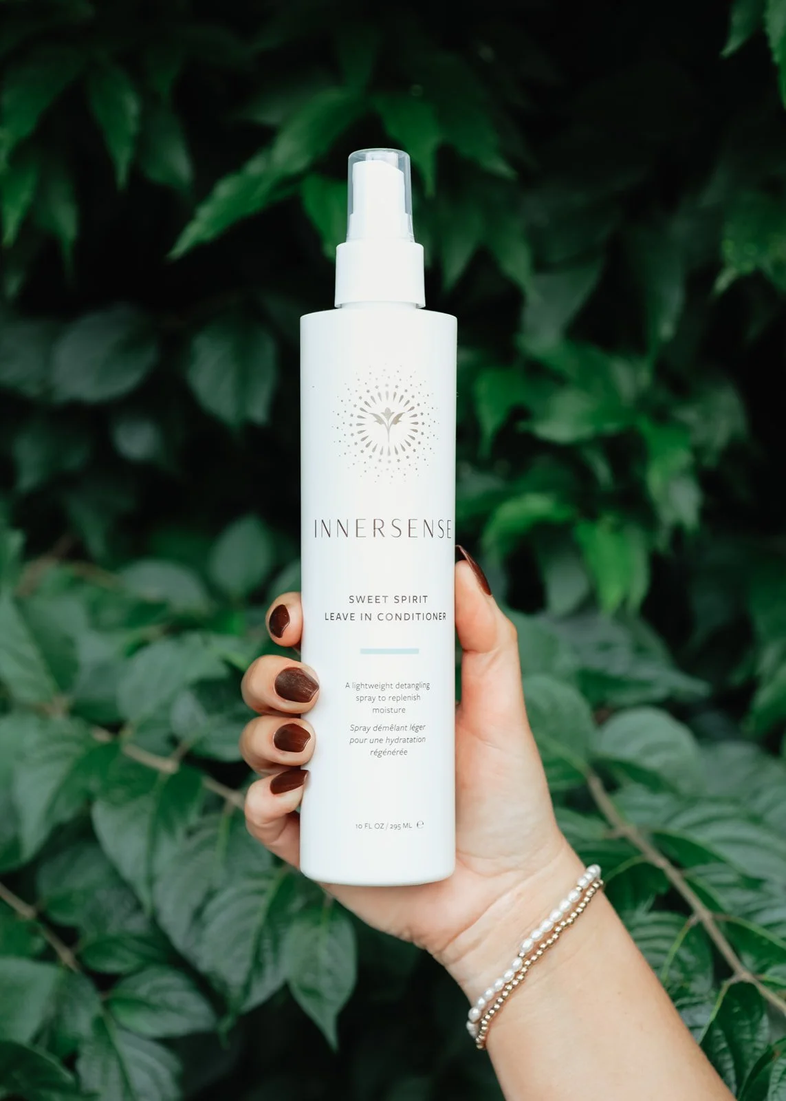 A hand holding a white bottle of INNERSENSE Sweet Spirit Leave-In Conditioner with a background of green leaves.