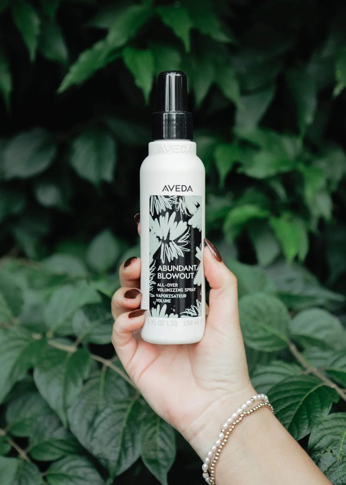 Hand holding a white bottle of Aveda Abundant Blowout volumizing spray with floral design label against green leafy background.