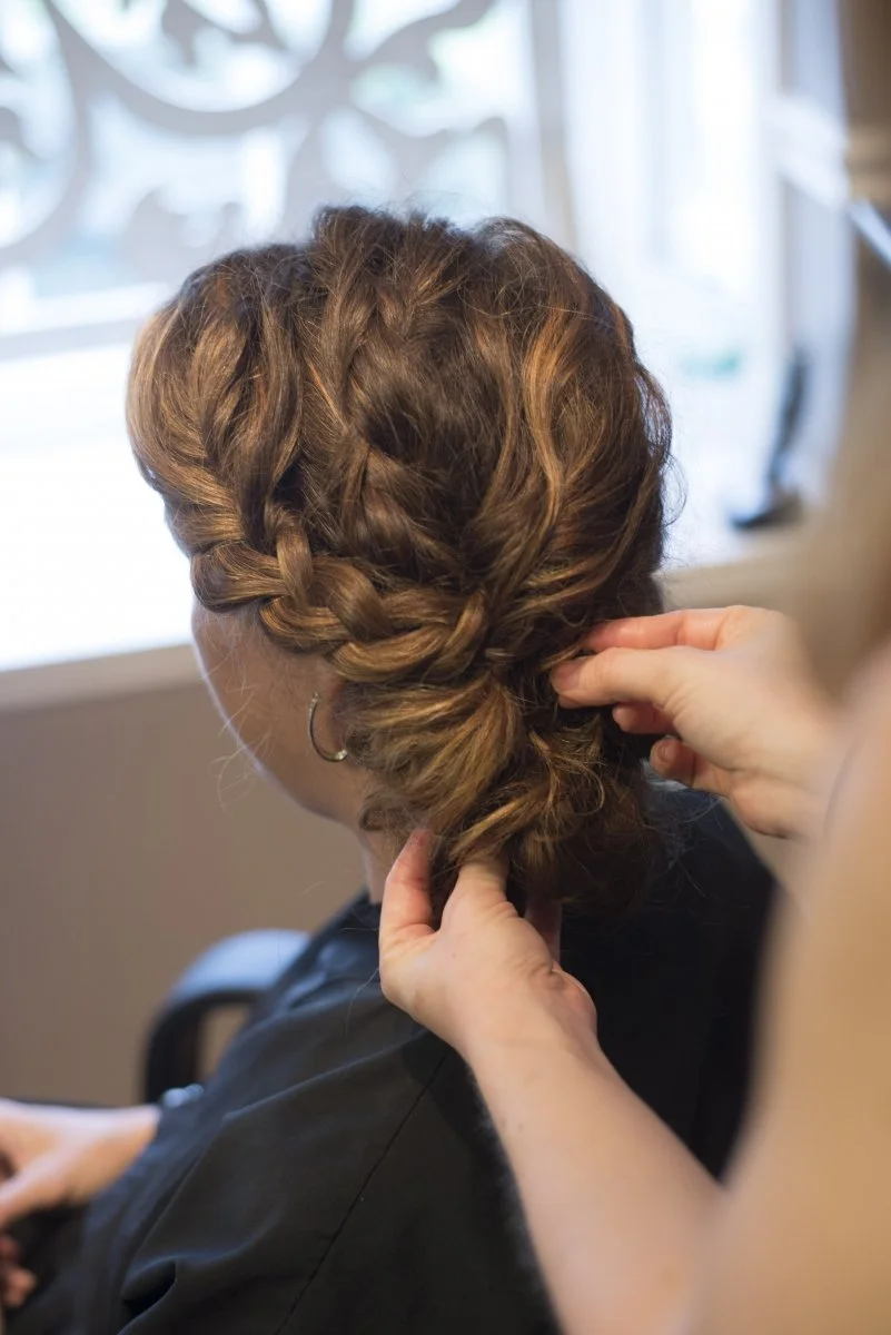 A person with short to medium length brown hair styled in a loose braid, sitting in a salon chair while a hairstylist works on their hair.