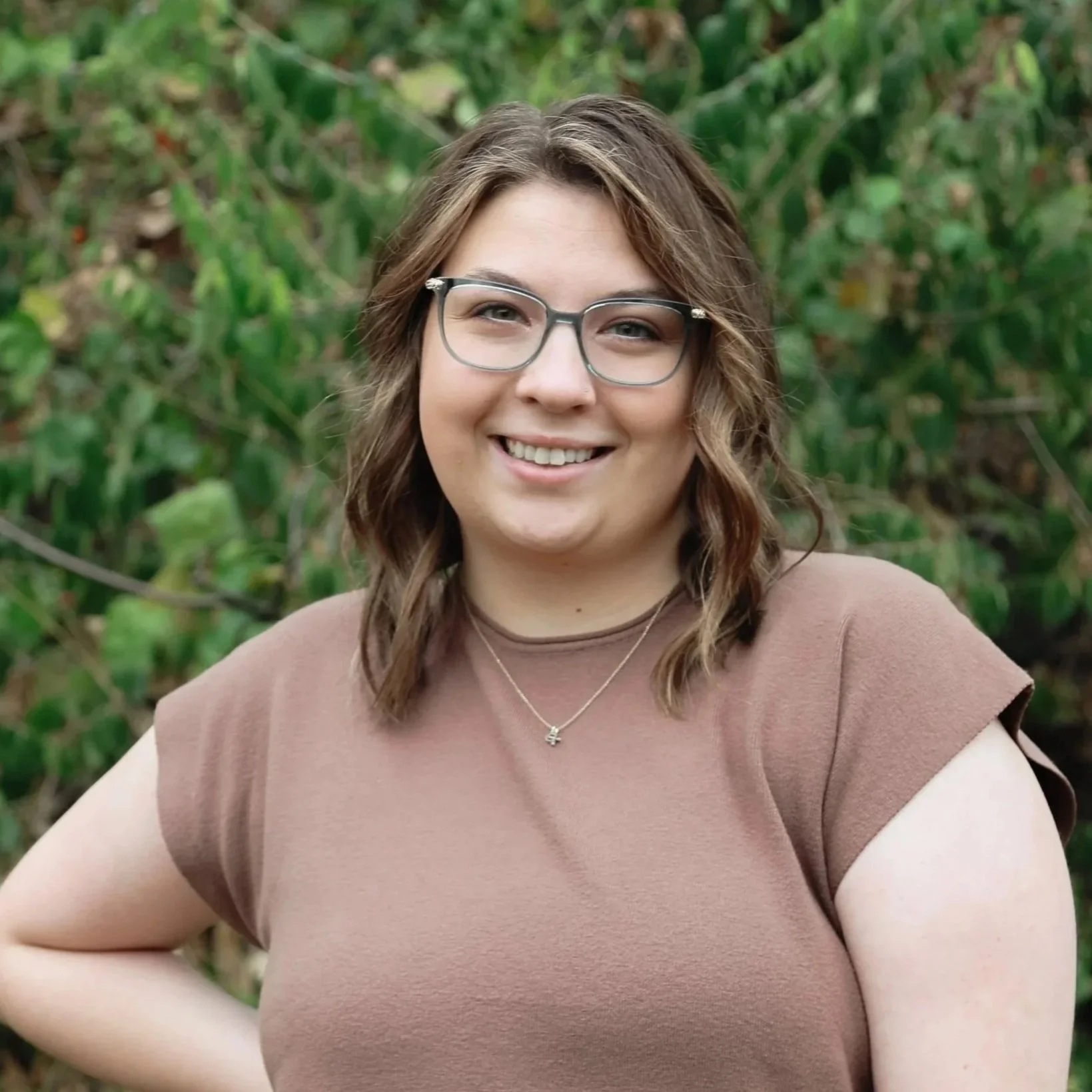 A woman smiling outdoors with greenery in the background. She has wavy brown hair, wears glasses, a tan shirt, and a silver necklace.