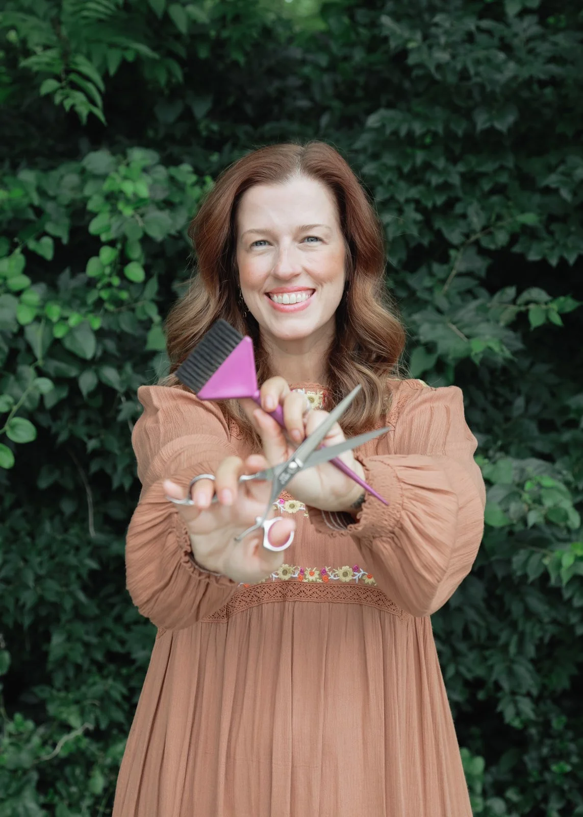 Woman with red hair smiling, holding scissors and a hairbrush, standing outdoors with green plants in the background.