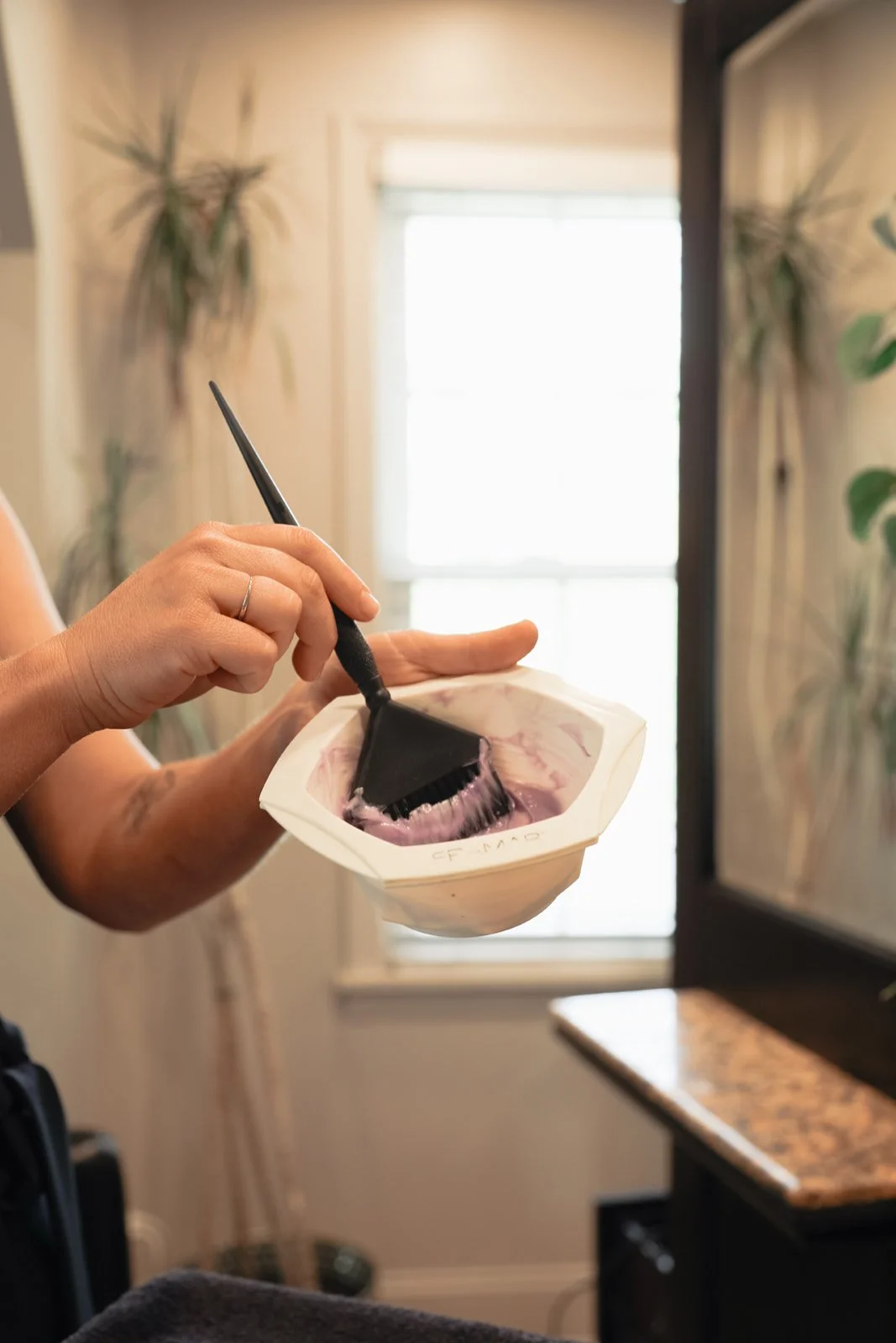 Person stirring a mixture in a bowl with a spatula in a well-lit room with houseplants and a window.