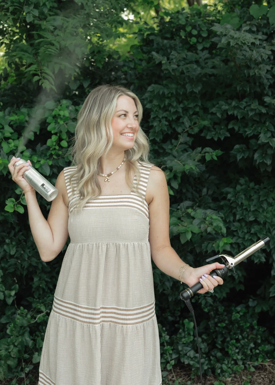 A young woman with wavy blonde hair outdoors, holding a can of hairspray in her right hand and a curling iron in her left, smiling and looking to her right, with green foliage in the background.