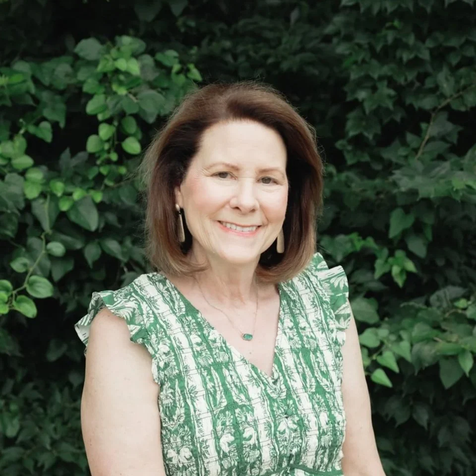 A middle-aged woman with shoulder-length brown hair, smiling, wearing a green and white patterned sleeveless top, earrings, and a necklace, standing outdoors in front of lush green foliage.
