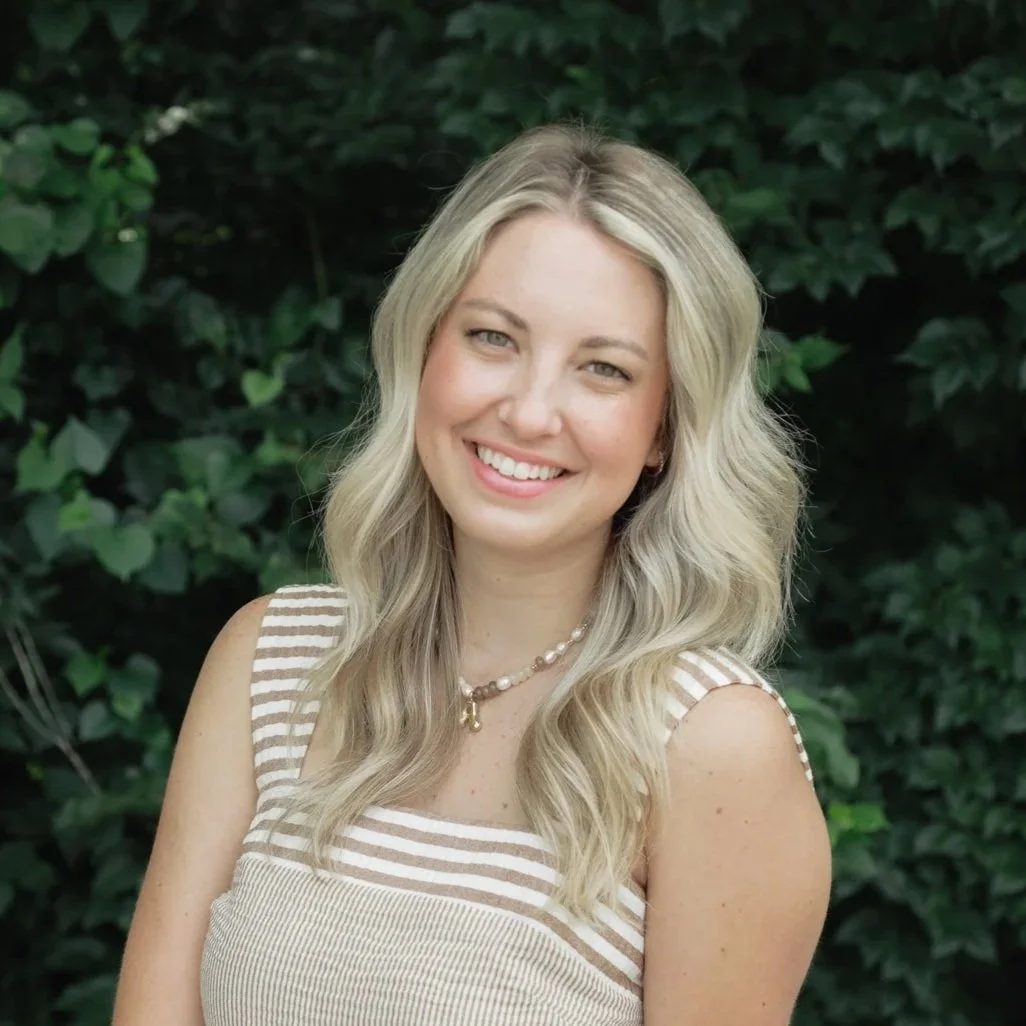 A young woman with long, wavy blonde hair smiling outdoors in front of green foliage, wearing a beige and white striped sleeveless top and a pearl necklace.
