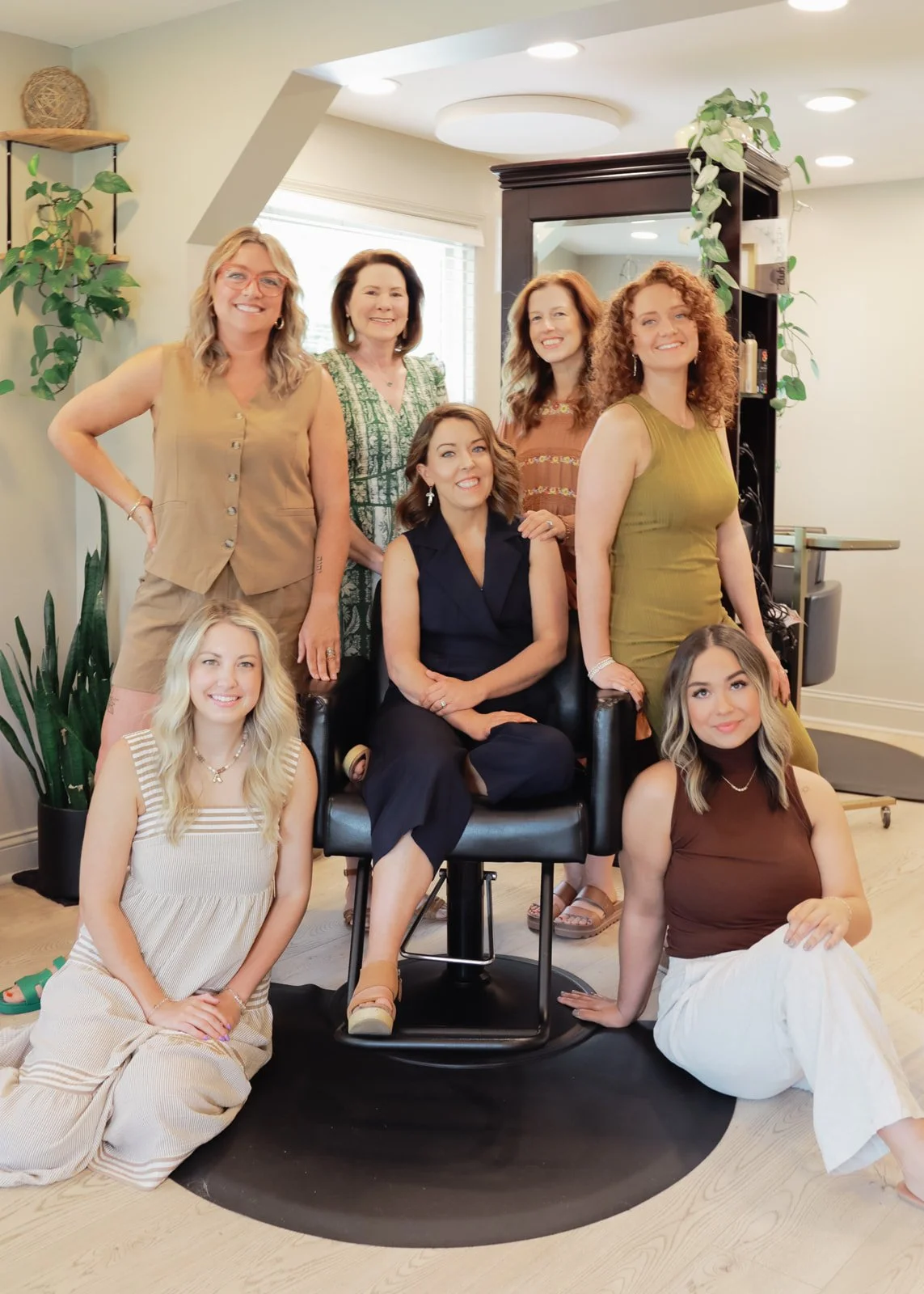 A group of nine women posed together in a salon or studio, smiling at the camera, with some seated and some standing, surrounded by mirrors, plants, and salon equipment.