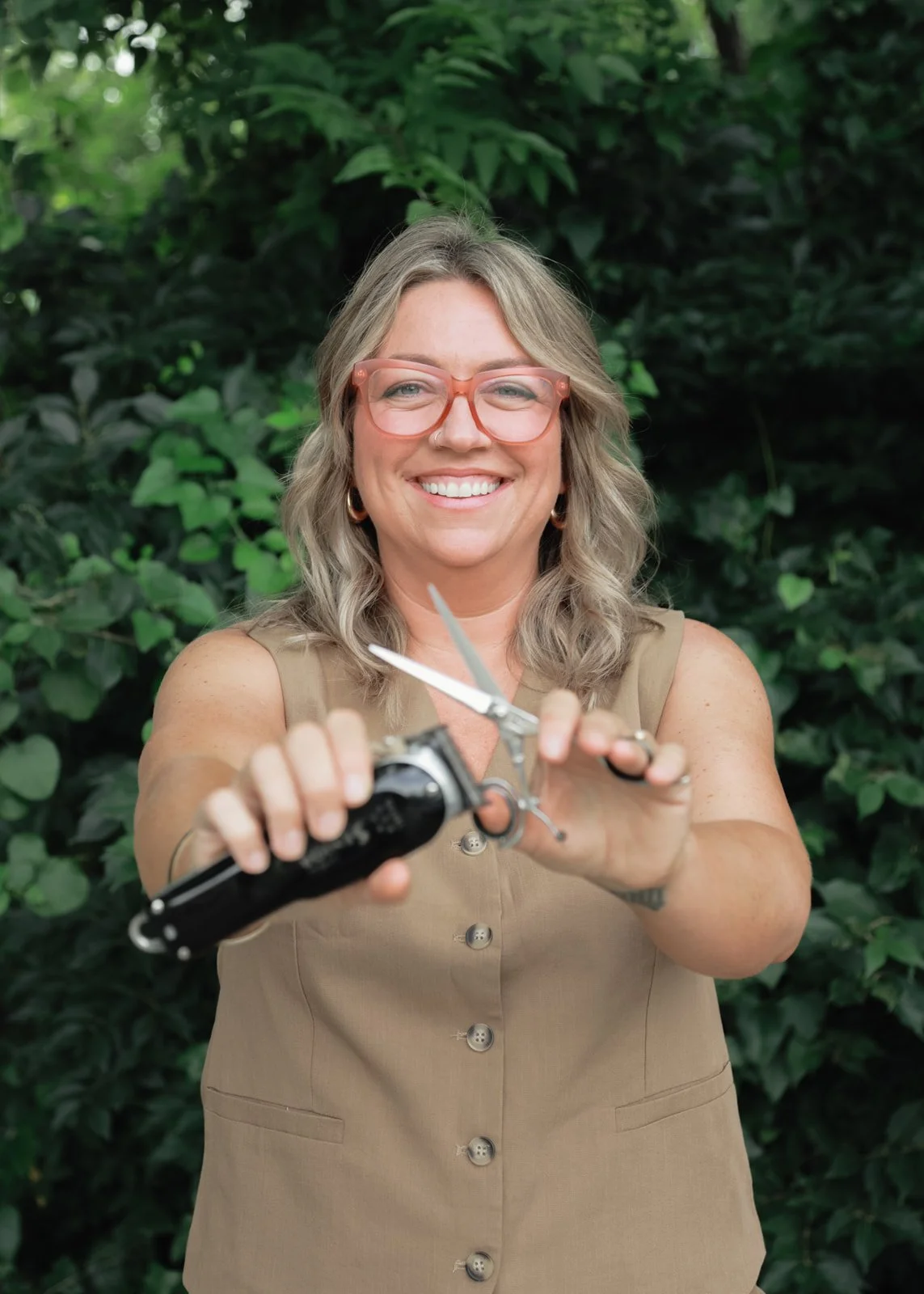 A woman with blonde hair, glasses, and a beige sleeveless dress, smiling and holding a pair of scissors towards the camera, in front of green leafy bushes.
