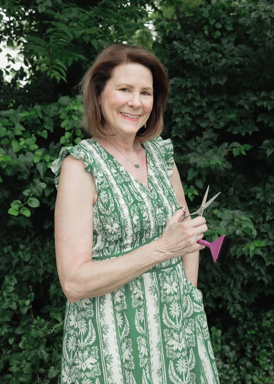 A woman with shoulder-length brown hair smiling and holding scissors and a pink hairbrush, standing outdoors in front of green leafy bushes. She is wearing a green and white floral dress and a necklace.