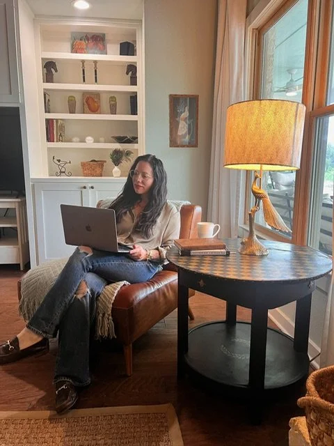 Woman sitting on a leather chair with a laptop, a lamp on a side table, and a window with curtains in a cozy living room.