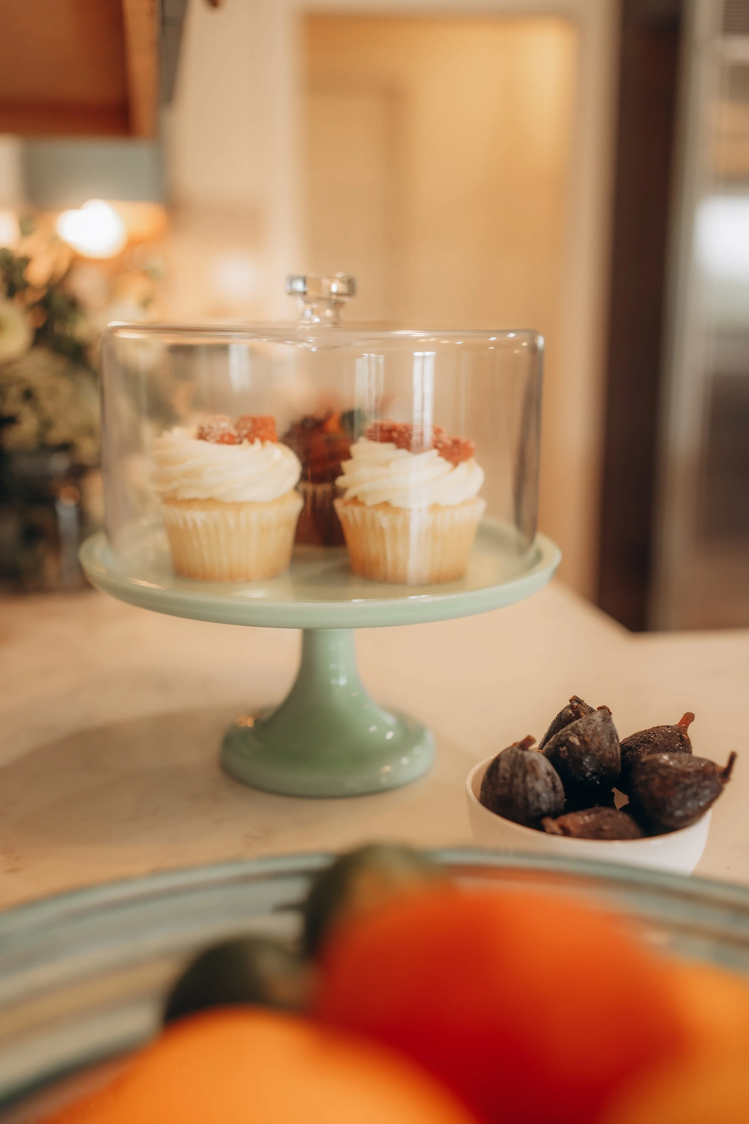 Close-up of cupcakes with white frosting and red toppings on a light green cake stand, with a blurred background of a lamp and holiday decorations, and a bowl of dark-colored sweets in the foreground.