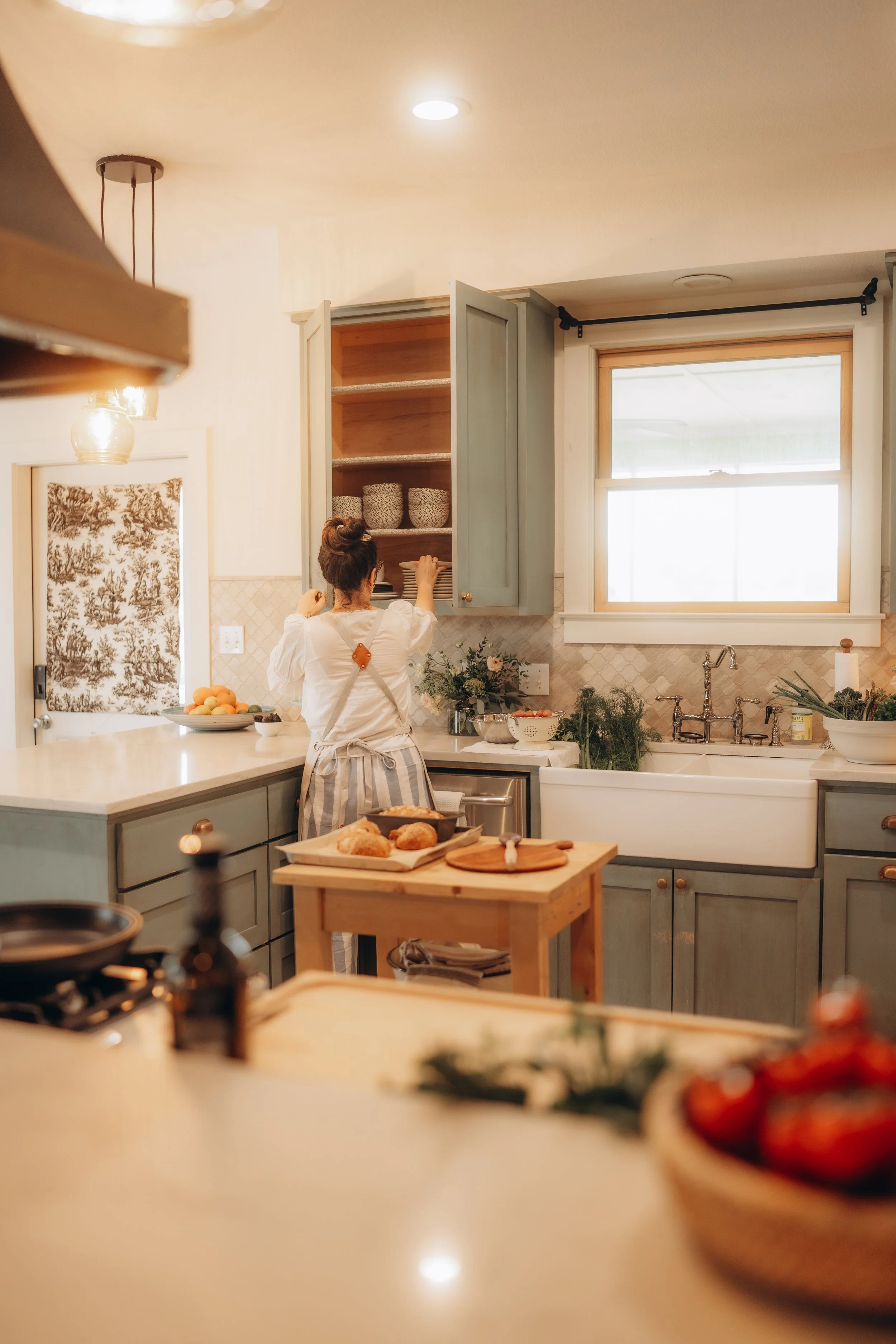 A woman in a white top and striped apron reaching into a kitchen cabinet, with a window above the sink, surrounded by plants and kitchen utensils, in a cozy, well-lit kitchen.
