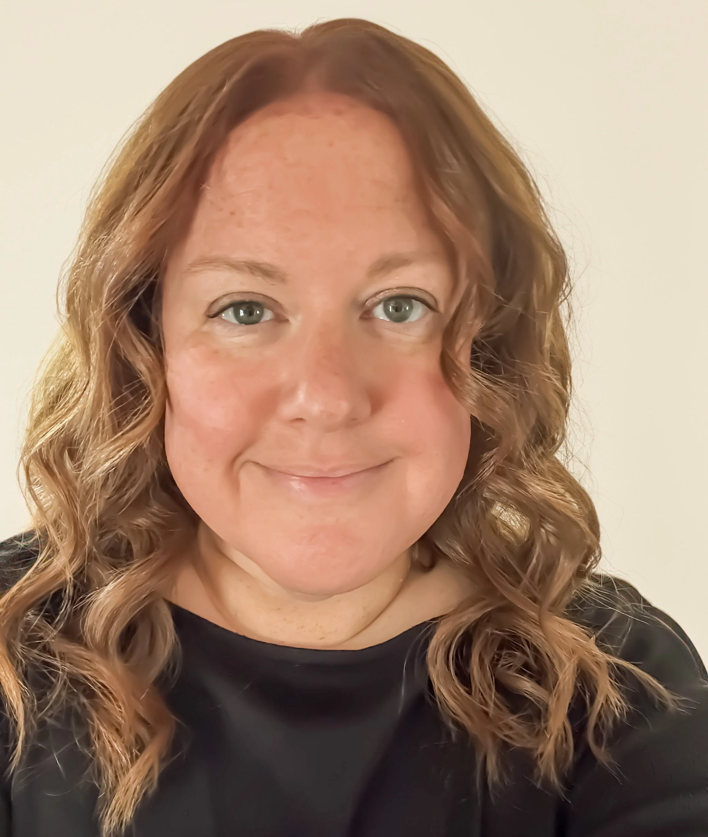 Close-up of a woman with light skin, wavy reddish-brown hair, green eyes, wearing a black top, smiling gently against a plain light-colored background.