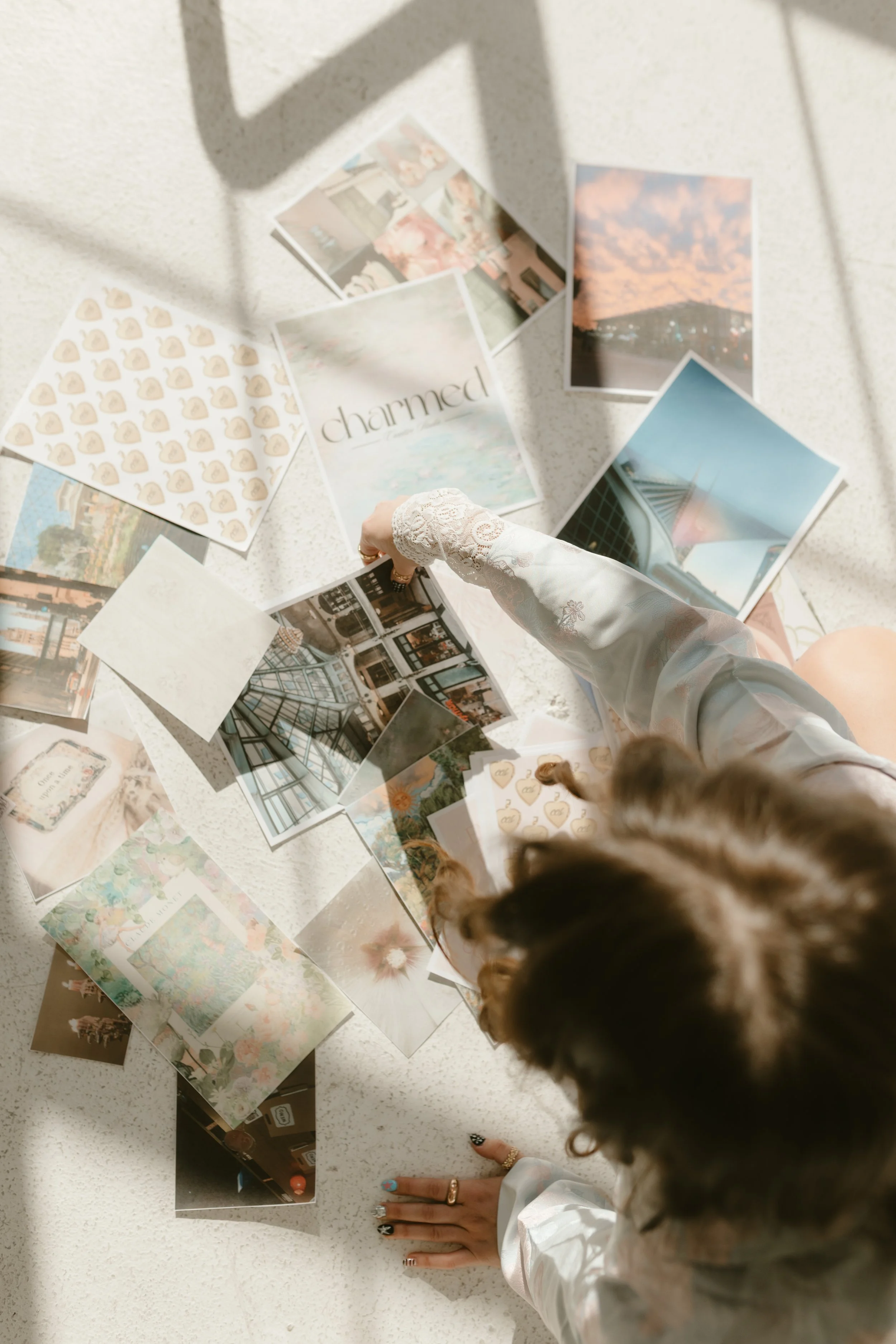 A person with curly hair, wearing a white lace long-sleeve top, is sitting at a white speckled table, pointing at various photographs and postcards spread out on the table, which include images of sky, buildings, and artwork. Shadows of a plant are cast on the table.