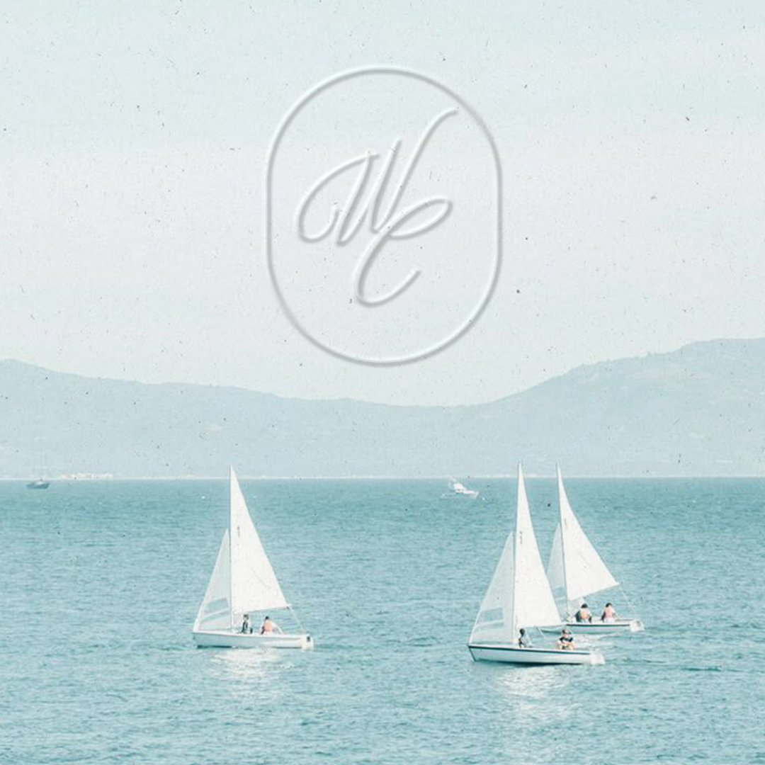 Three sailboats with white sails floating on calm blue water, distant mountain range and a few small boats in the background.