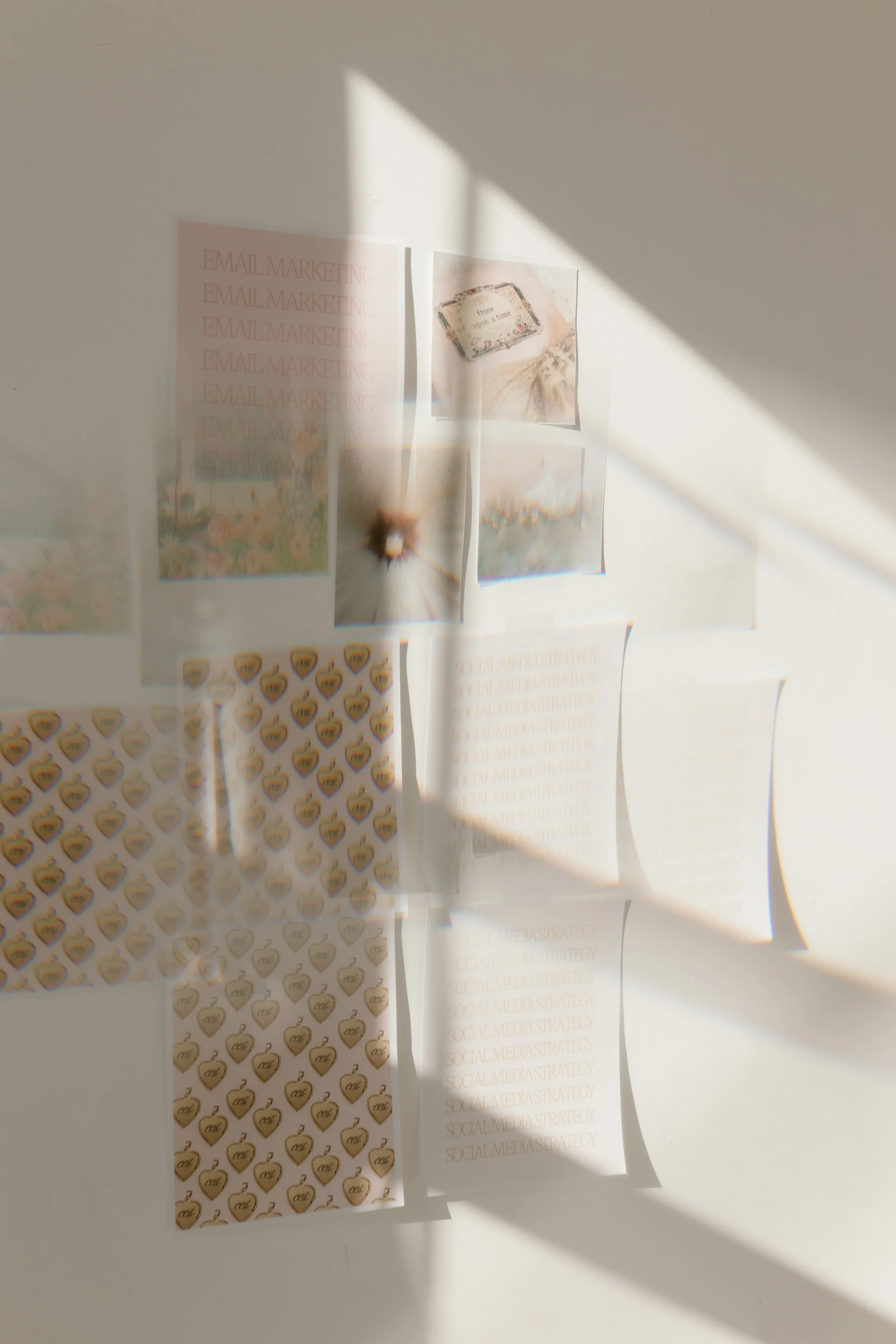 A white wall with several sheets of paper and images taped to it, including some text that says 'Email Marketing' and 'Social Media Strategy', illuminated by sunlight and shadows.