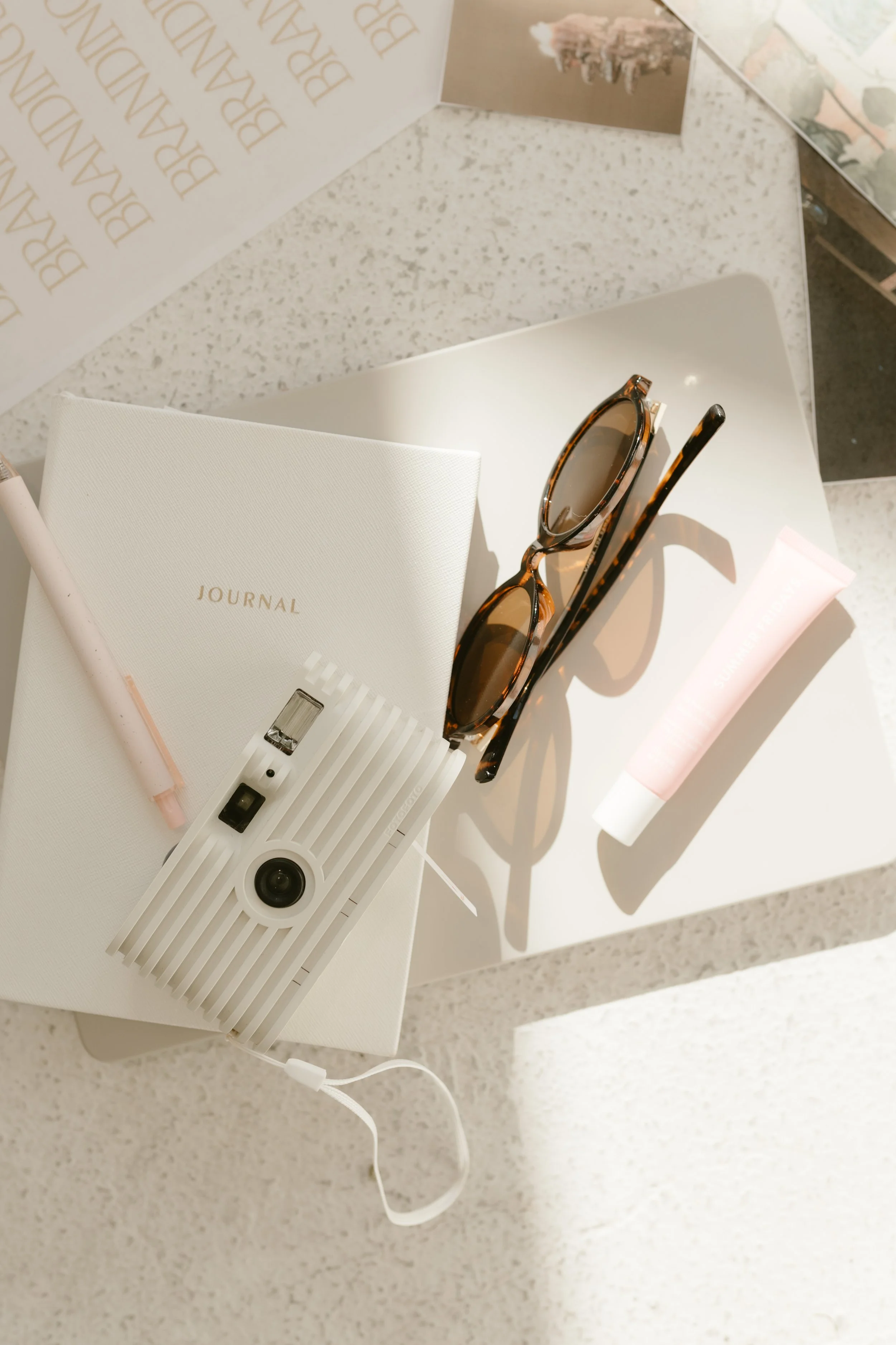 A white table holding a pair of tortoiseshell sunglasses, a white journal, a pink and white pen, a pink tube of sunscreen, and a white projector on a speckled white countertop.