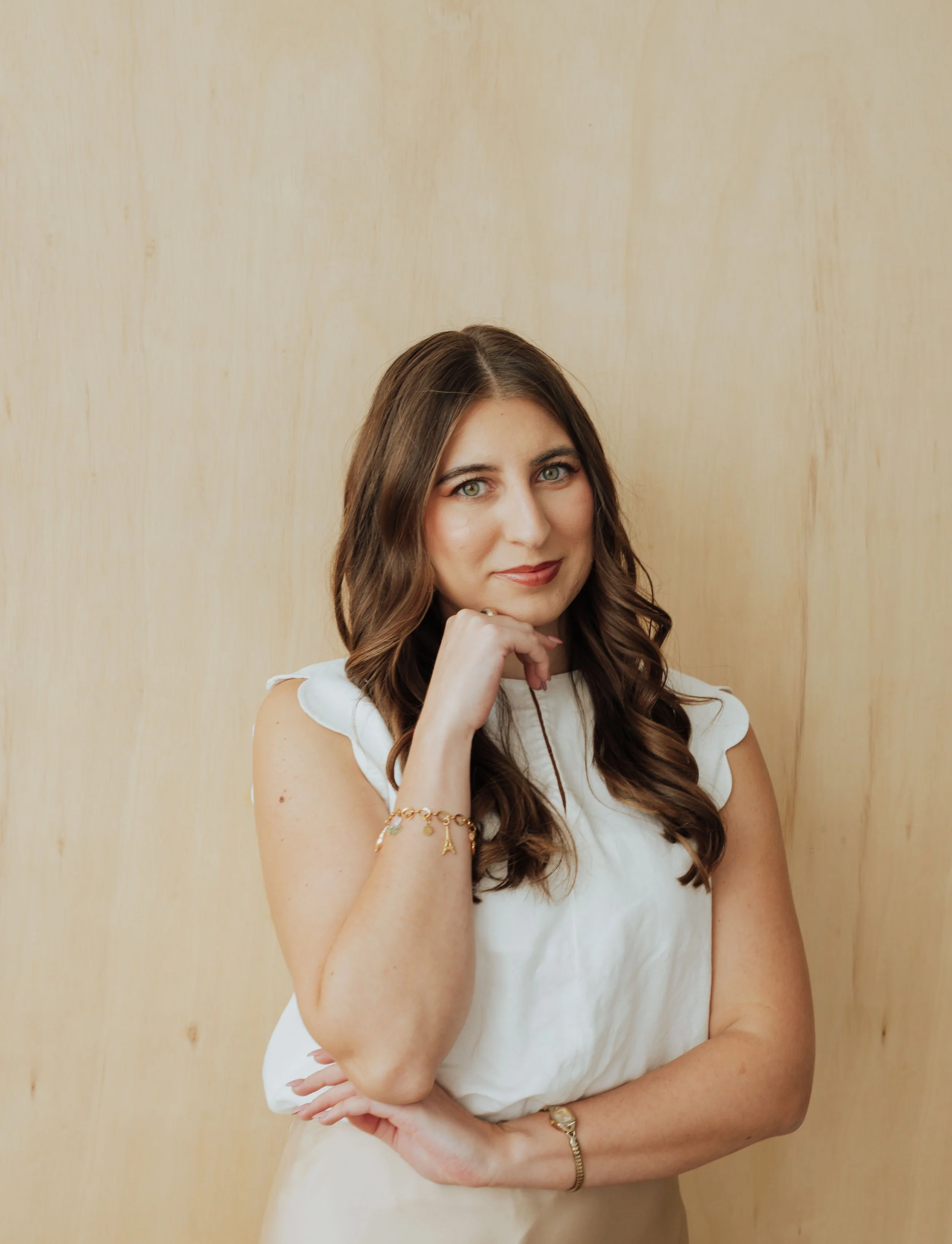 A woman with long brown hair and blue eyes, dressed in a white sleeveless top, standing against a wooden wall, resting her chin on her left hand, and smiling softly.