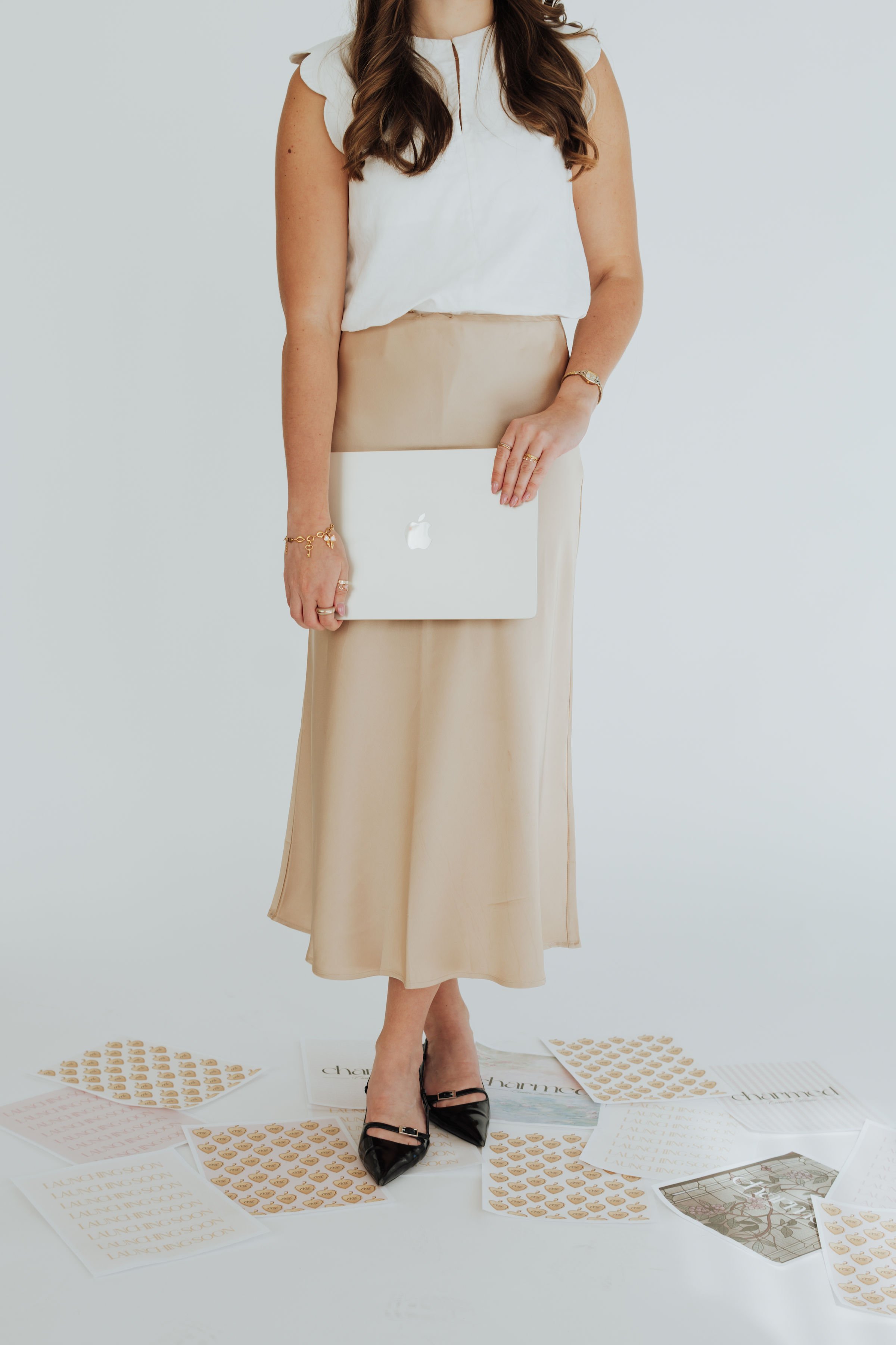 A woman in a white top and beige skirt holding a silver laptop, standing among sheets of paper with printing, on a white floor against a plain light background.