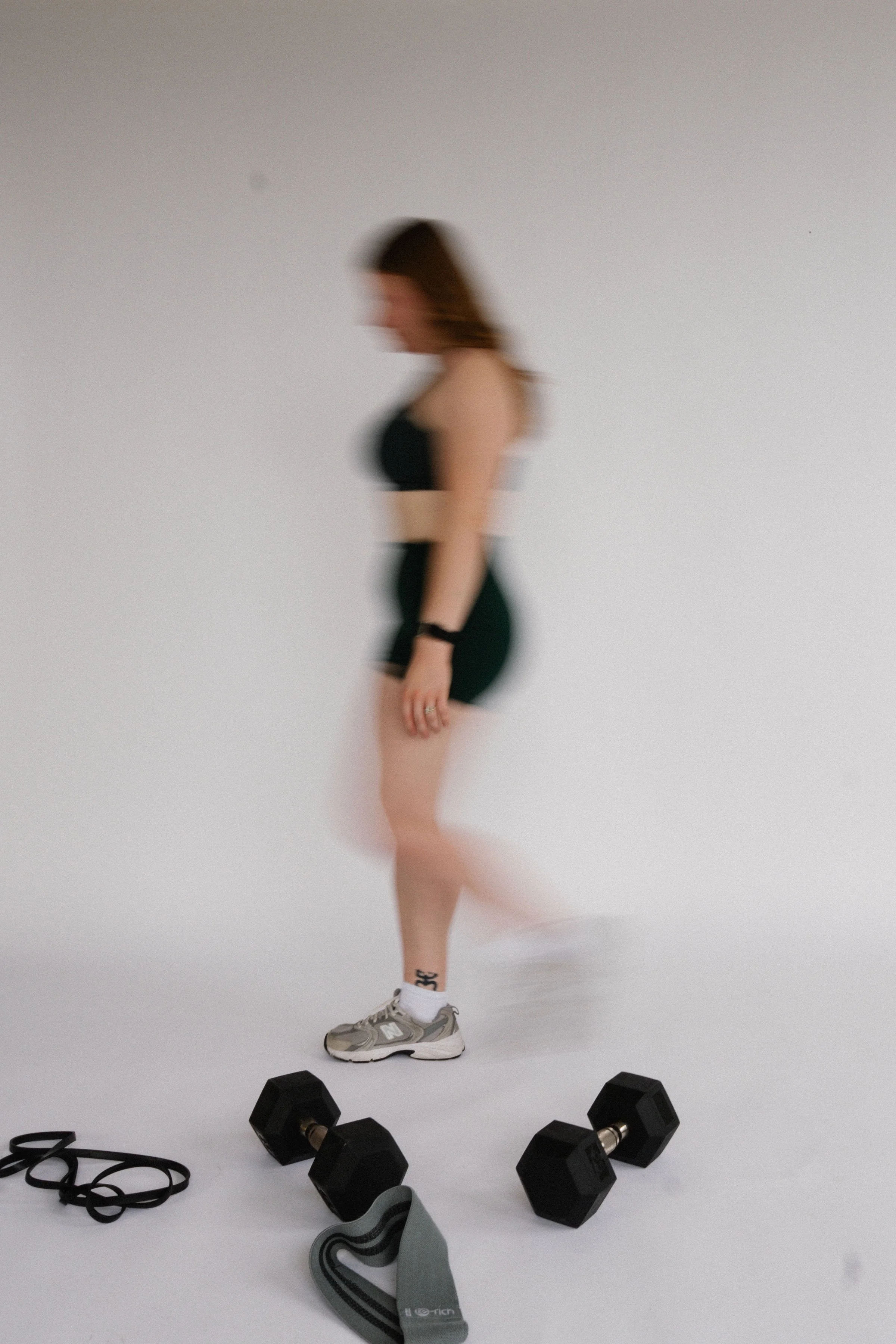 A woman in black workout clothes and sneakers stands on a plain background, with dumbbells, resistance bands, and fitness accessories around her feet.
