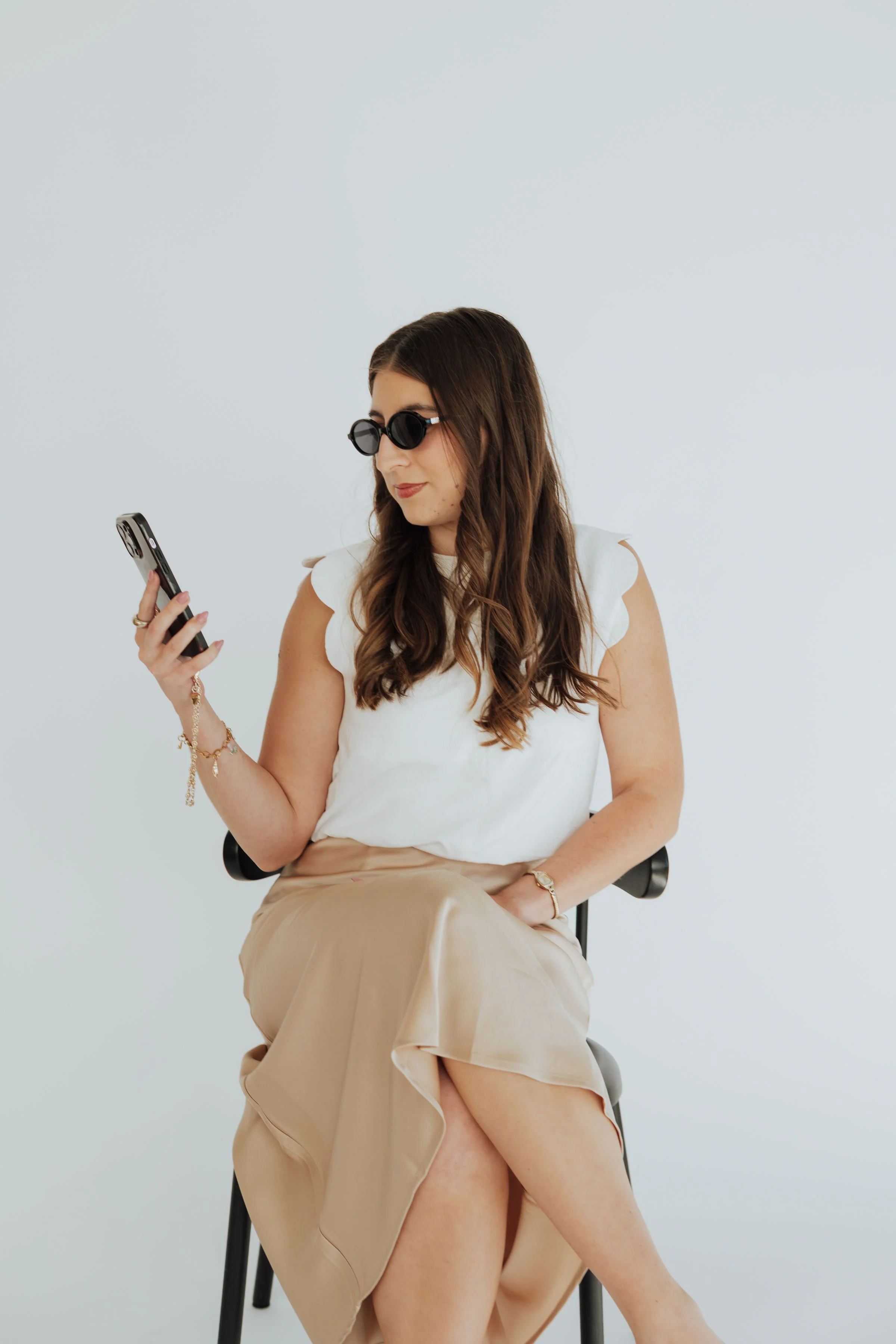 A woman wearing black sunglasses, a white top, and beige skirt sits on a black chair against a plain white background, looking at her phone.
