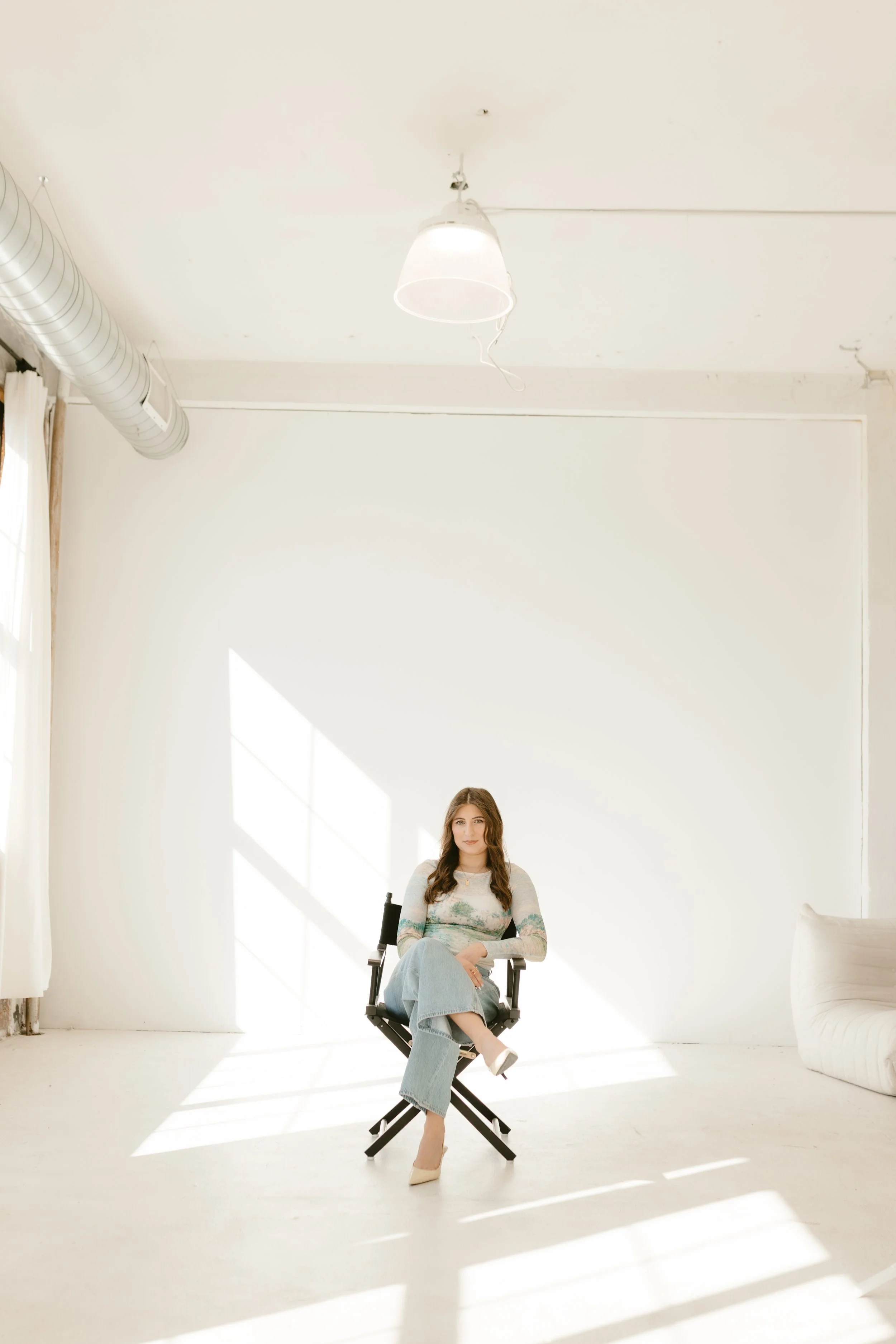 A woman sitting on a director's chair in a bright, minimally decorated room, with sunlight casting shadows through a large window. She has long brown hair and is wearing a casual patterned top and light blue jeans.