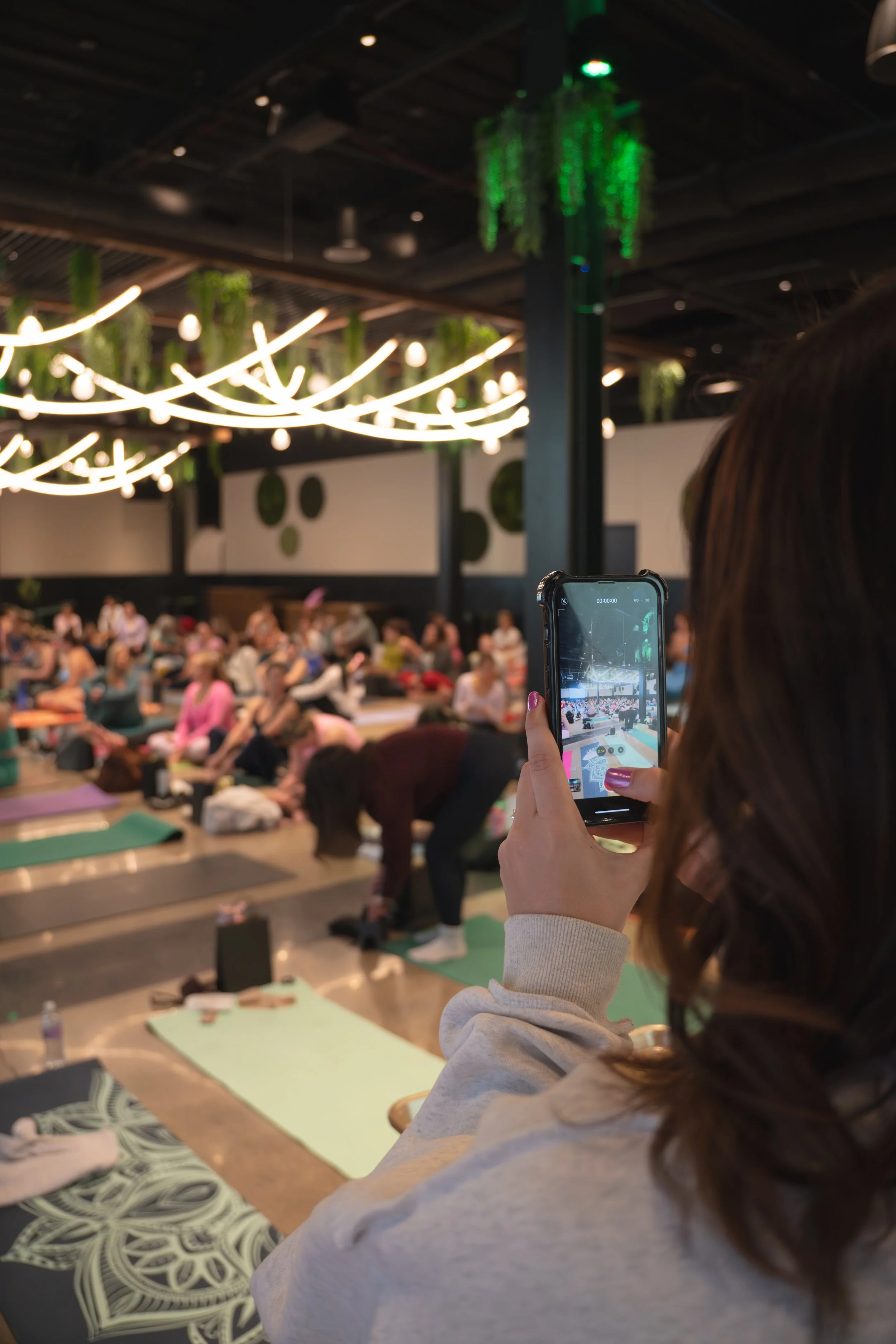 A woman holding a smartphone and taking a picture of a yoga class in a spacious, modern studio with green and white decor, hanging lights, and a large group of people practicing yoga on mats.