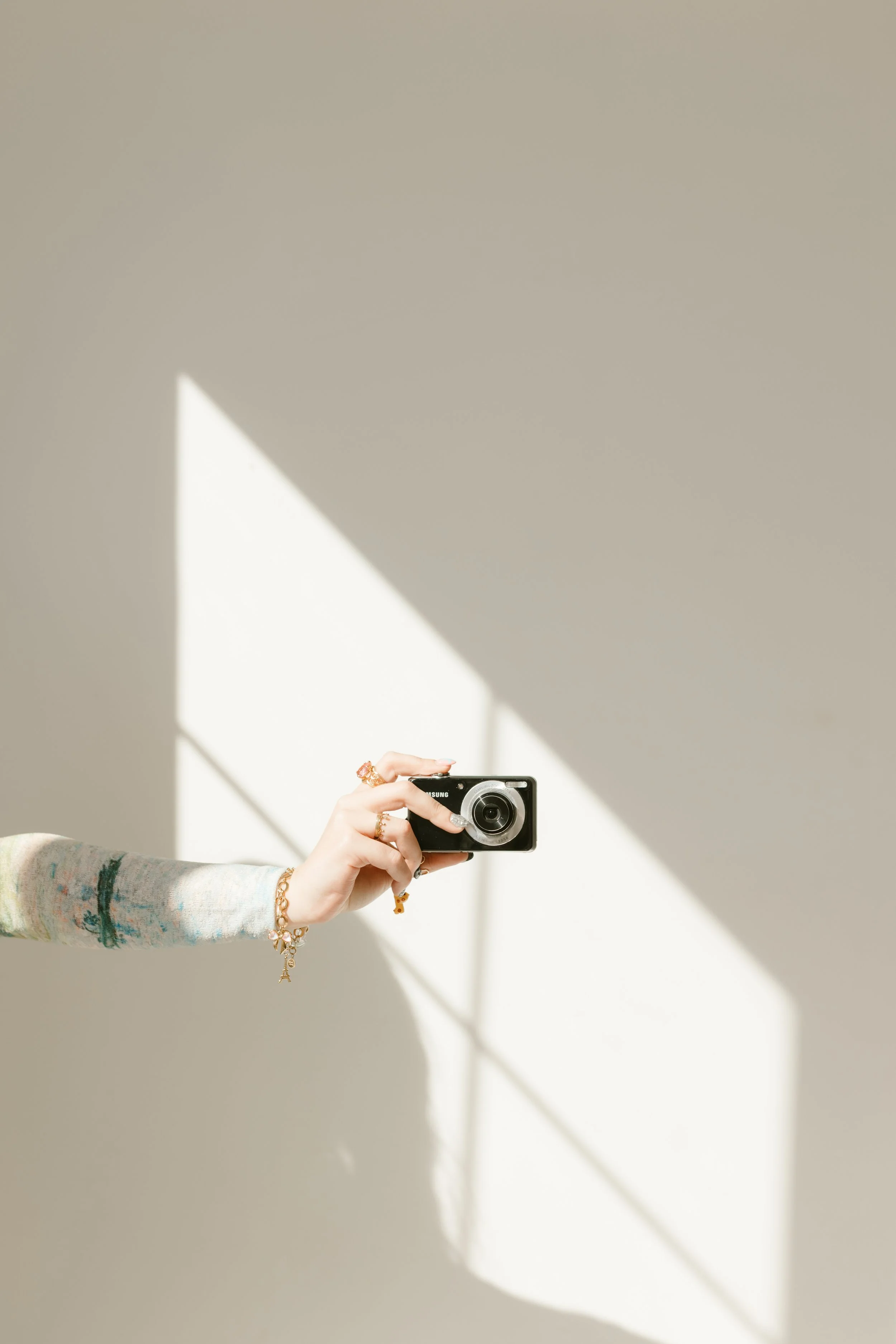 Person taking a photo with a camera, wearing jewelry and a pastel-colored sweater, against a beige wall with sunlight casting shadows.