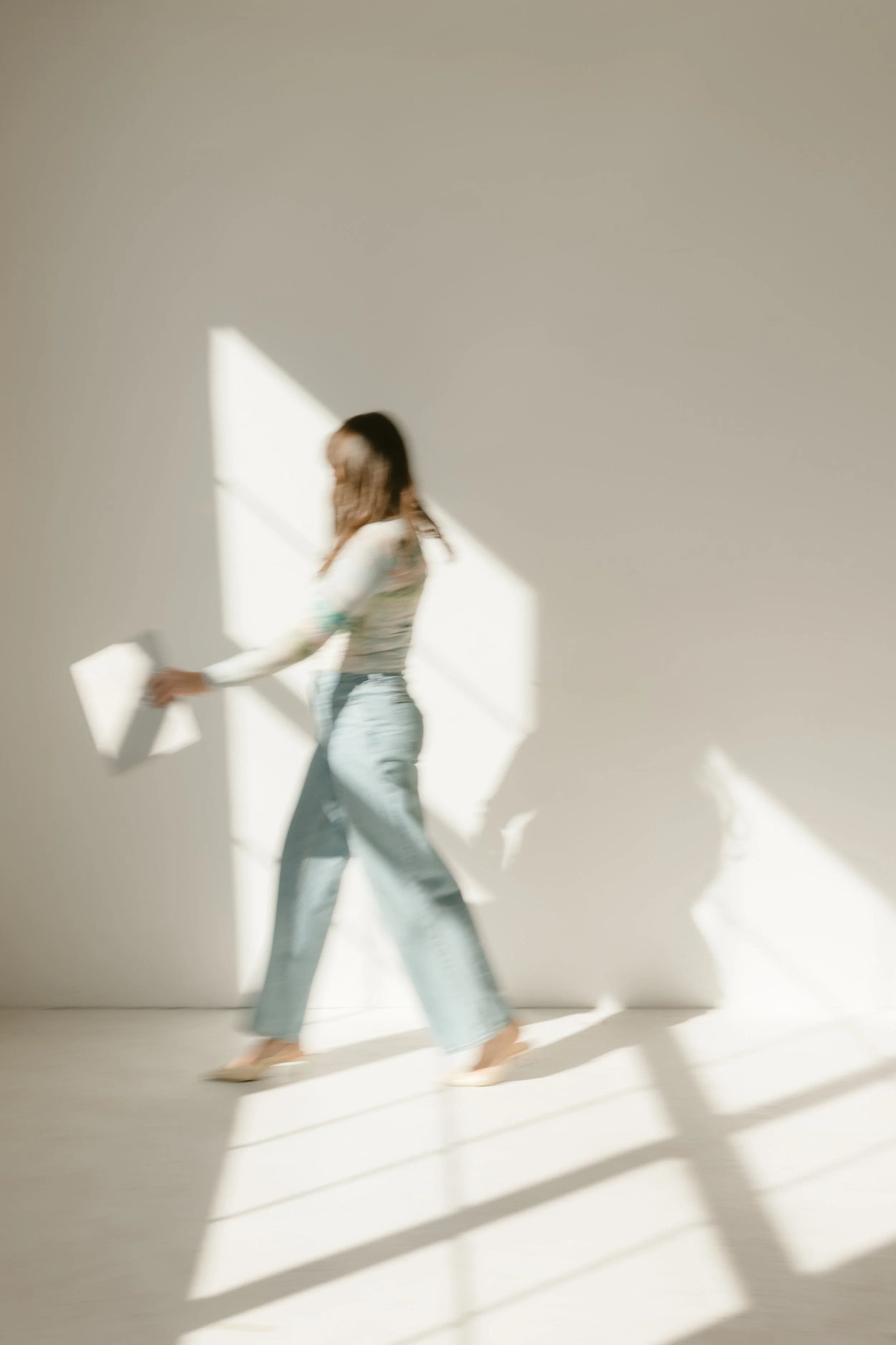 A woman with long hair, wearing a long-sleeved shirt and wide-leg jeans, walking barefoot on a light-colored floor next to a plain wall, with sunlight casting shadows on the wall.
