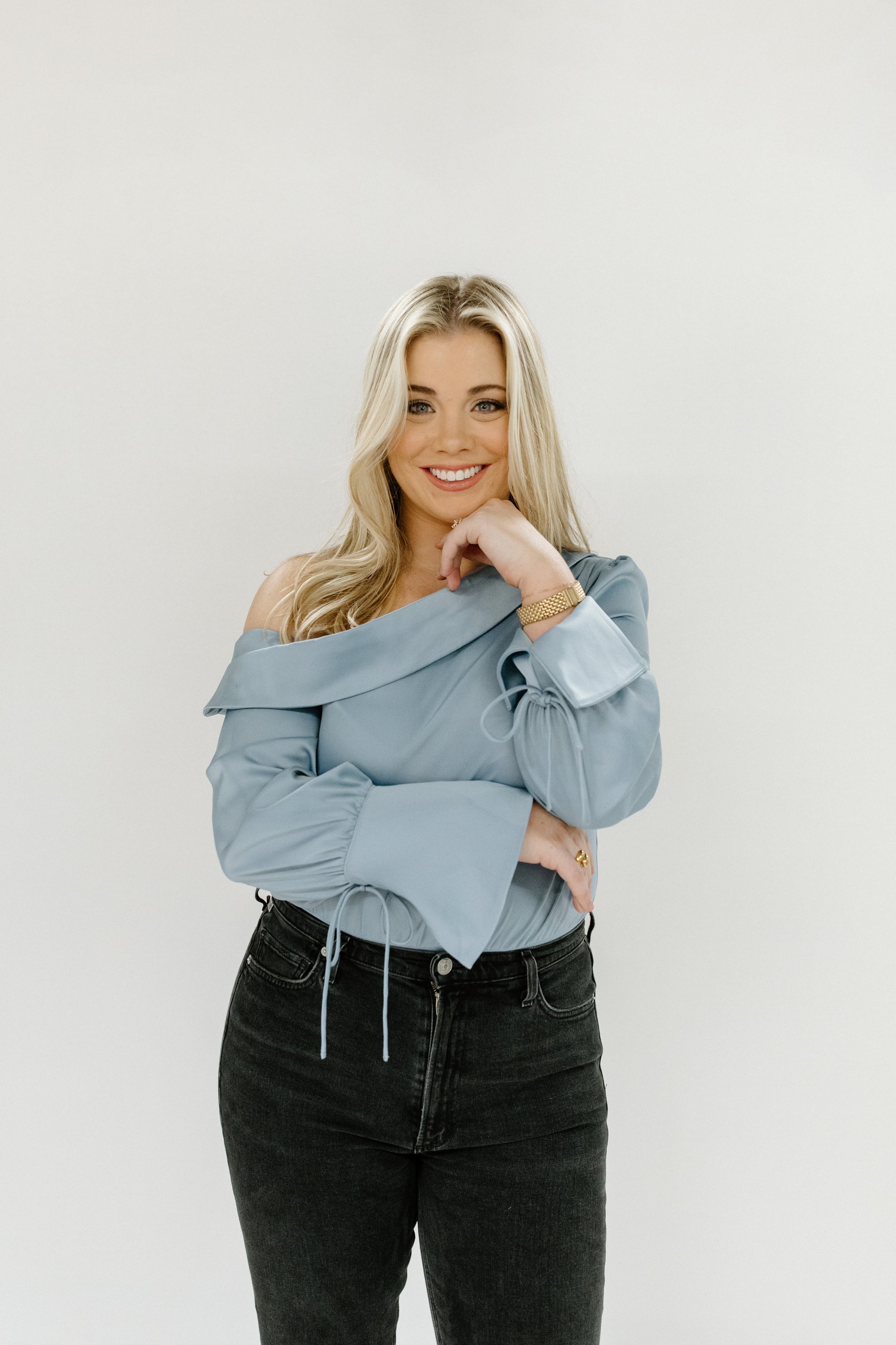 A woman with blonde hair smiling, wearing a light blue off-shoulder blouse and black jeans, standing against a plain white background.