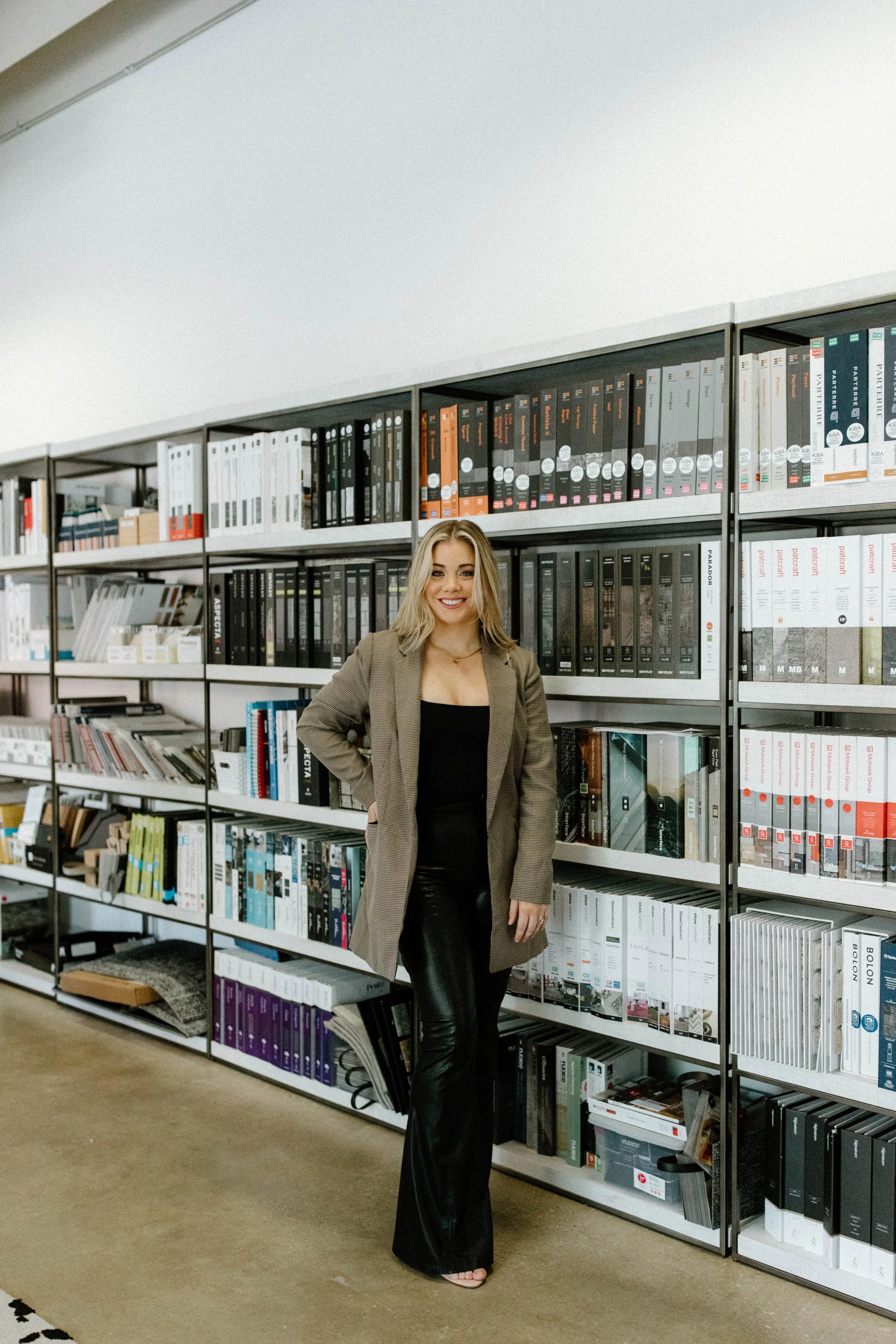 A woman with blonde hair, wearing a brown plaid coat, black top, and black leather pants, stands in front of bookshelves filled with binders and books in an office or library setting.