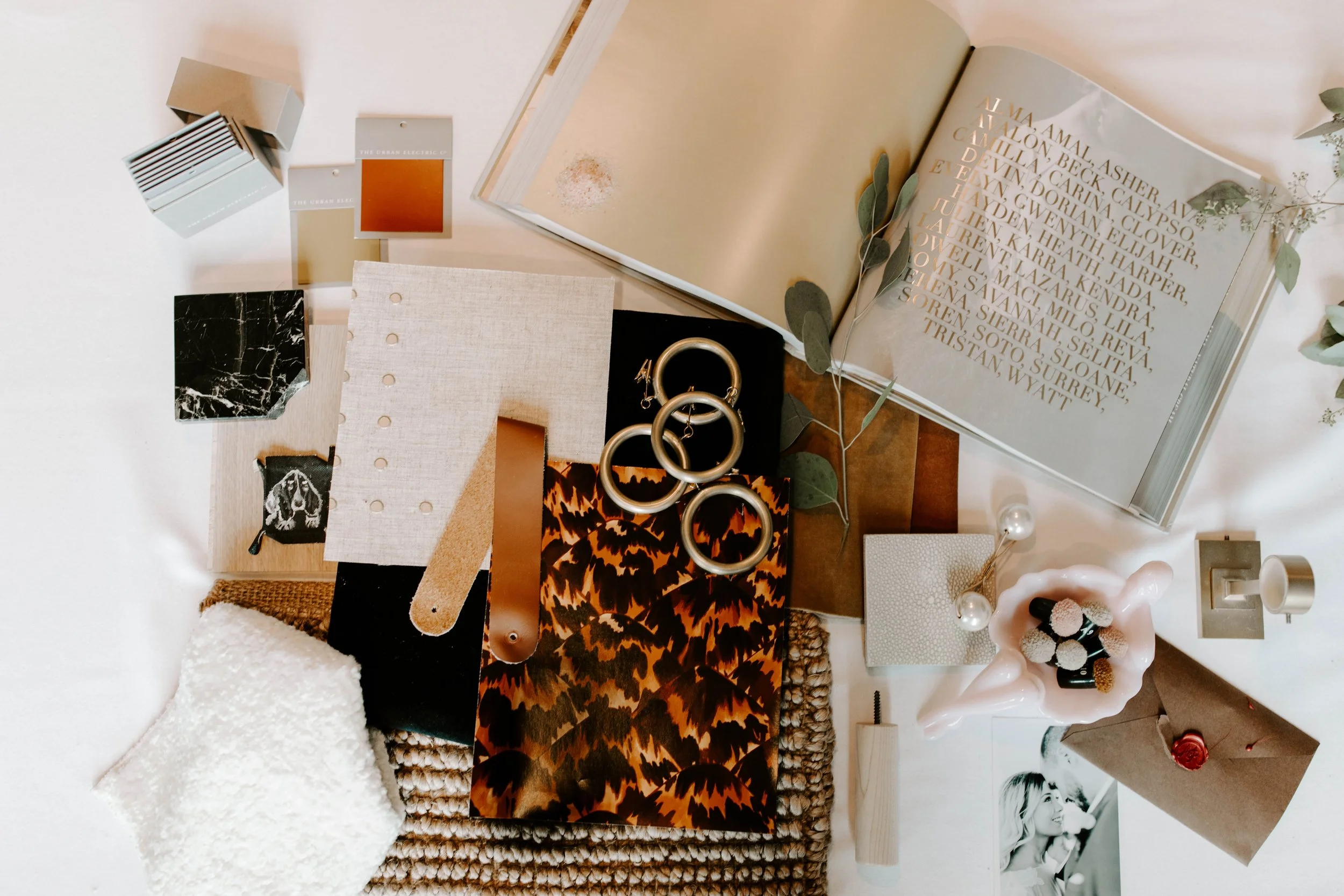 A flat lay of various design and decor materials, including paint samples, fabric swatches, jewelry, an open book with gold text, plant branches, a black marble tile, and a pink ceramic sculpture, arranged on a white surface.