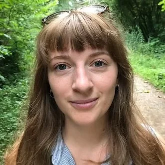 A young woman with long brown hair and bangs, wearing sunglasses on her head, smiling outdoors with greenery and a dirt path in the background.