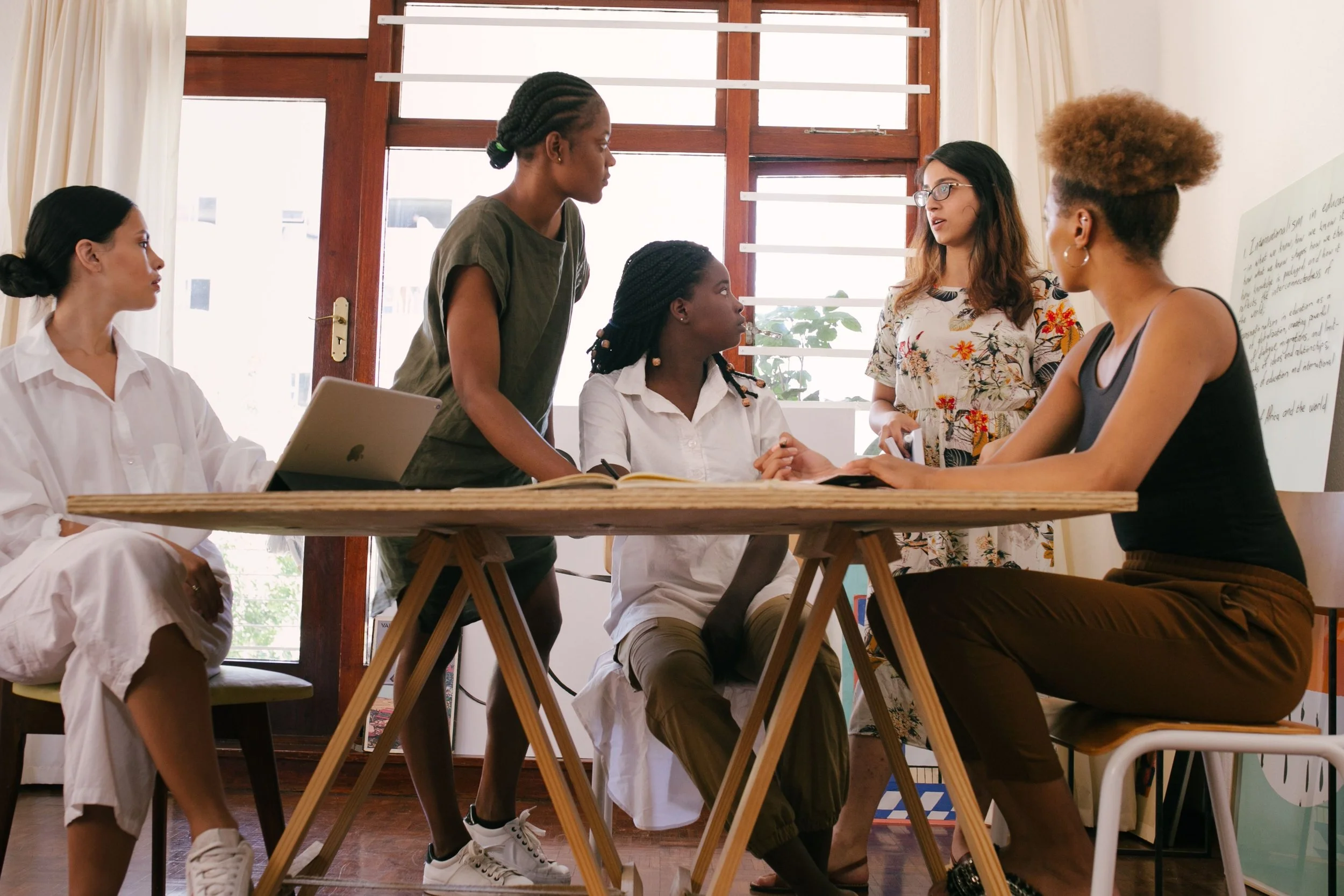 Group of five women having a discussion in a room with large windows and white curtains.