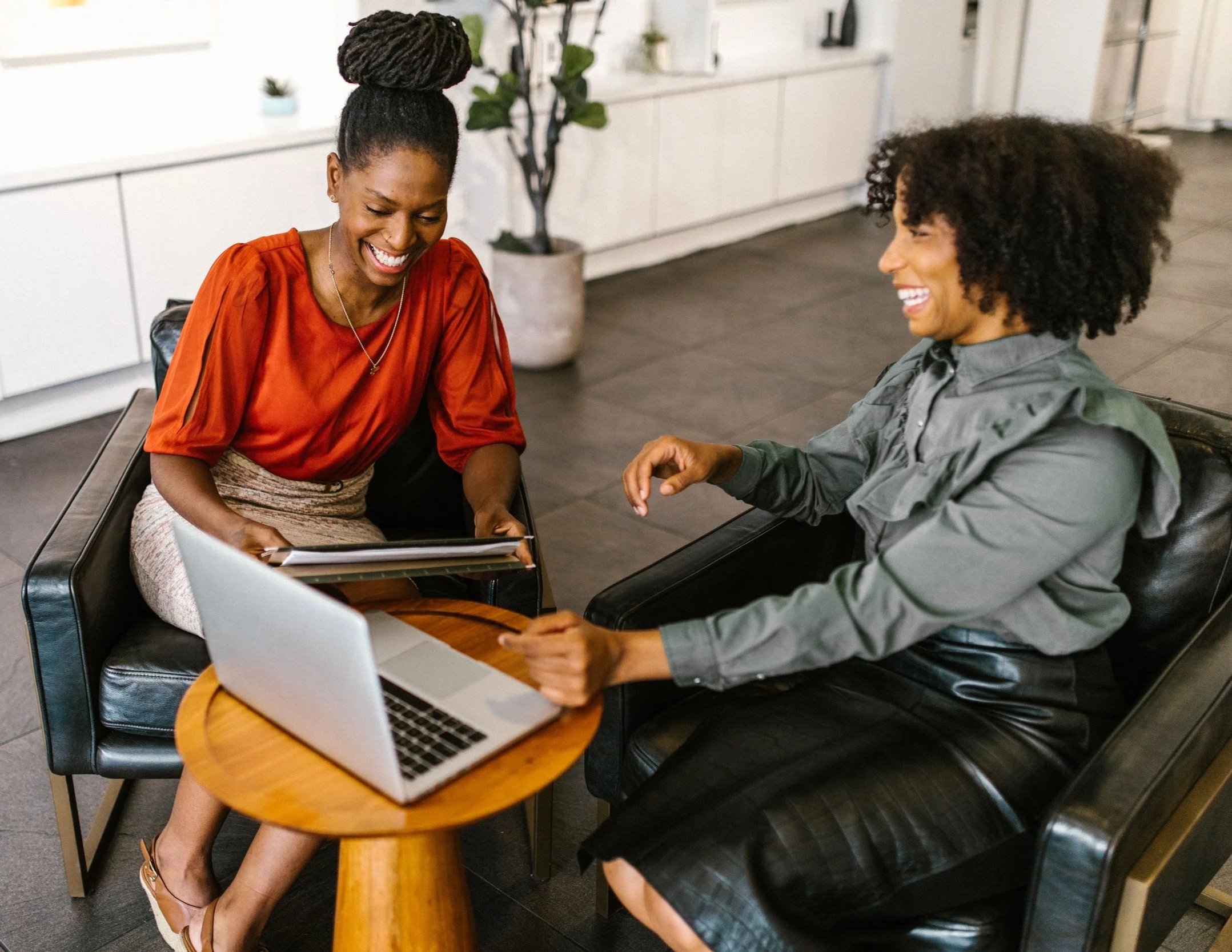 Two women sitting at a small round wooden table, laughing and looking at a laptop. One woman has braided hair styled in an updo and is wearing an orange top, while the other woman has curly hair and is wearing a gray jacket.