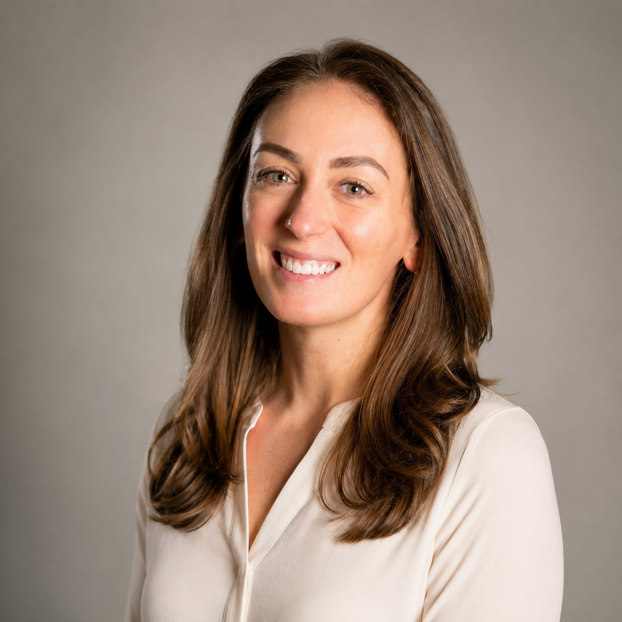 A woman with long brown hair smiling at the camera, wearing a cream-colored blouse, against a plain gray background.