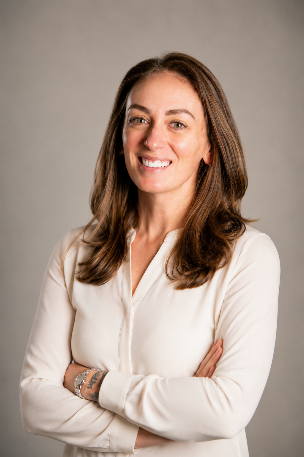 Portrait of a smiling woman with long wavy brown hair, wearing a cream-colored blouse, arms crossed, against a plain gray background.