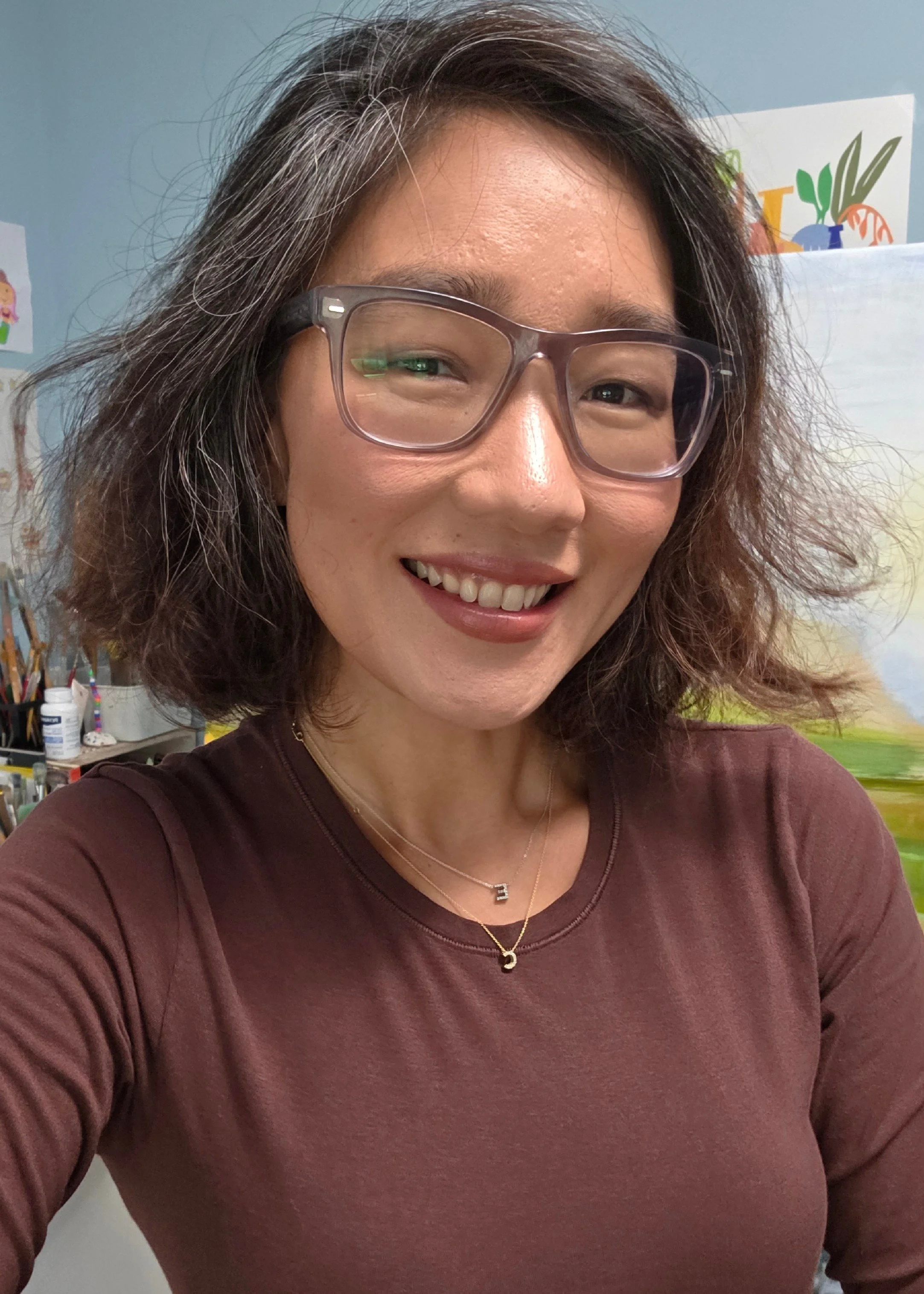 A woman with shoulder-length dark hair wearing glasses, a maroon top, and layered necklaces smiling in an indoor setting with artworks and art supplies in the background.