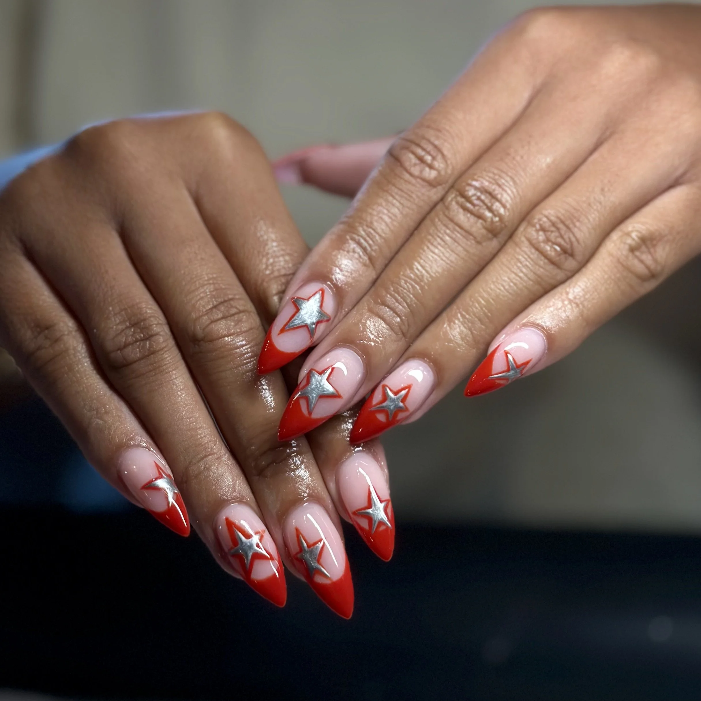 Close-up of a person's hand with star-designed red, white, and gray nail art, holding the fingertips of another hand against a dark background.