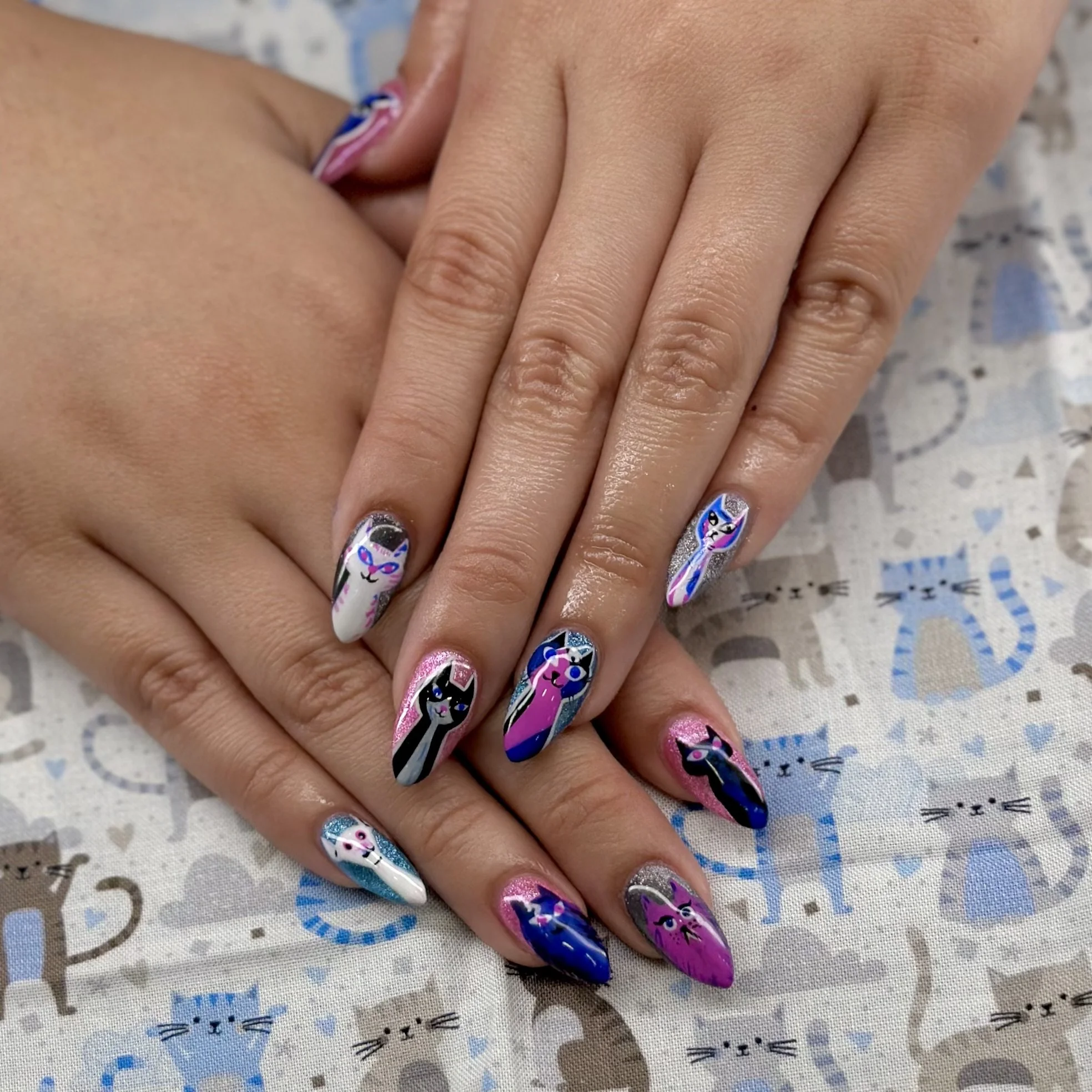 Close-up of a person's hand with long, decorated manicured nails featuring unicorn and rainbow designs, resting on a surface with a printed pattern of cats and hearts.