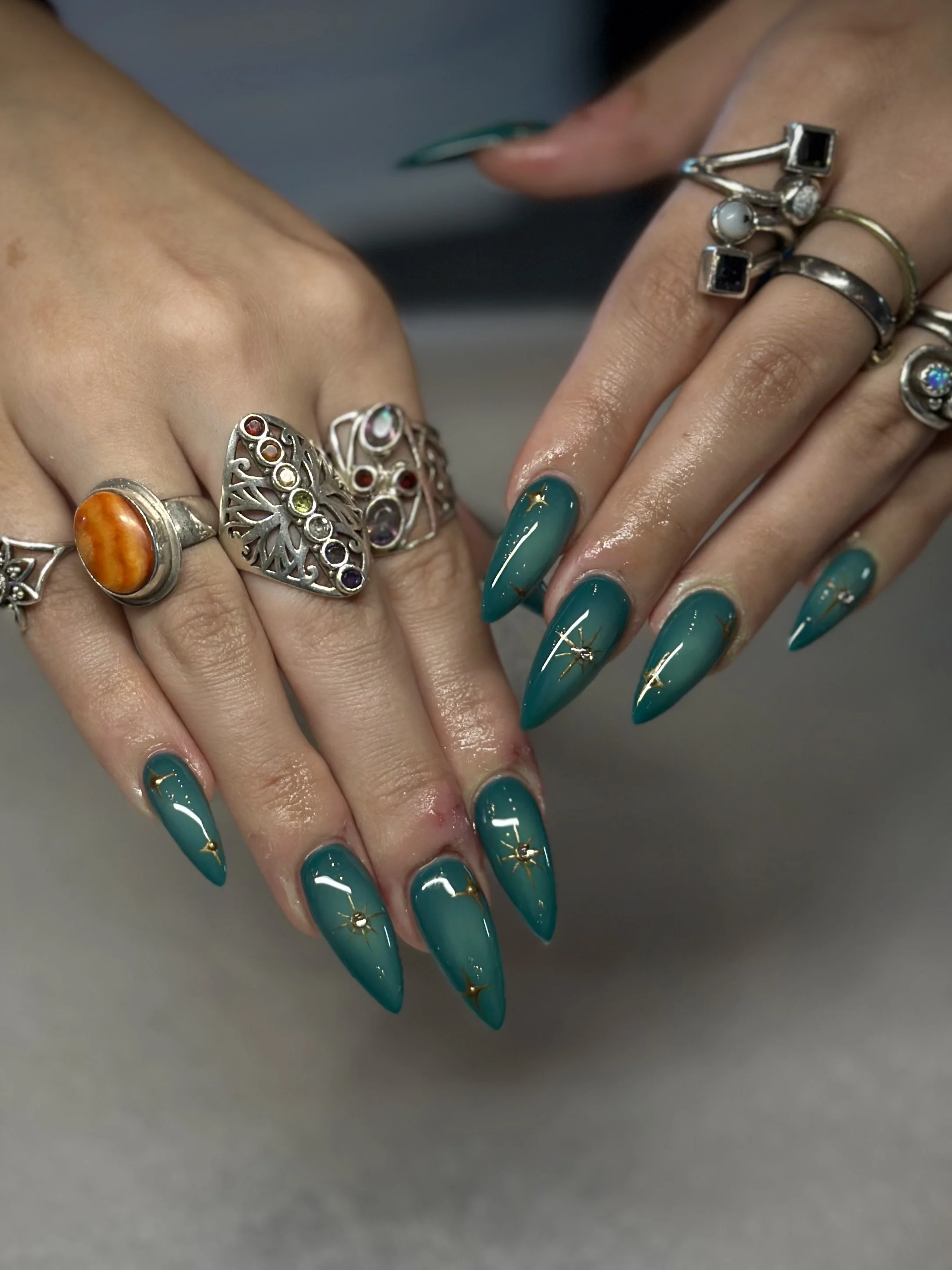 Close-up of hands with teal nail polish, gold star and stud decorations, and multiple silver rings with various gemstones and designs.