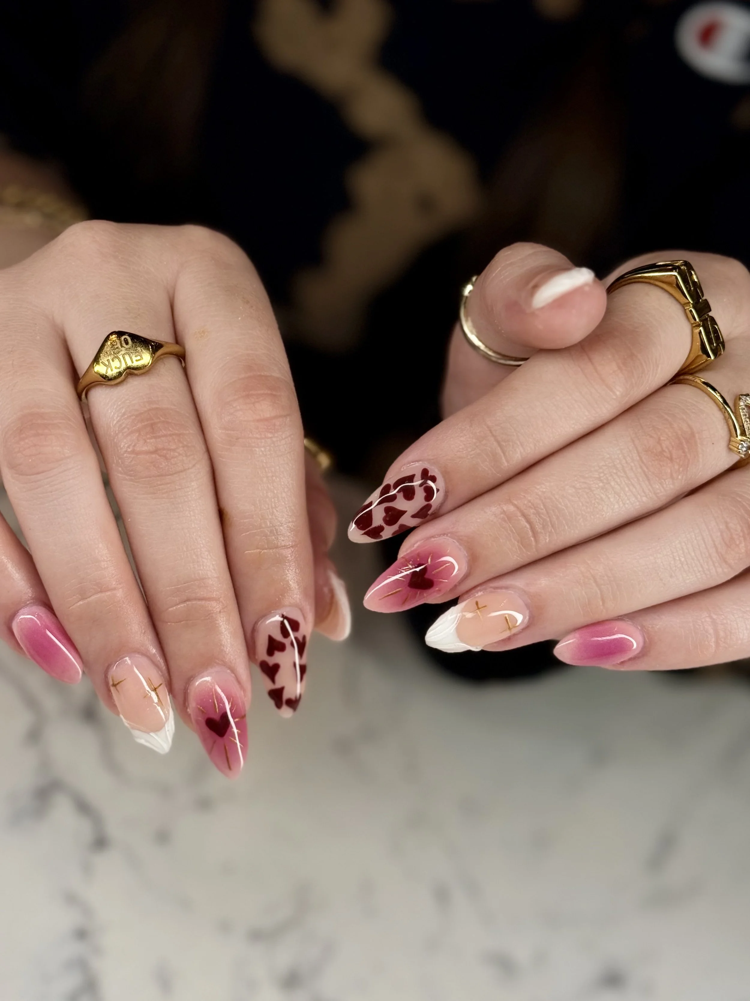 Close-up of person’s hands with Valentine's Day themed nail art and gold rings, some rings engraved with words, person's fingernails decorated with hearts and words.
