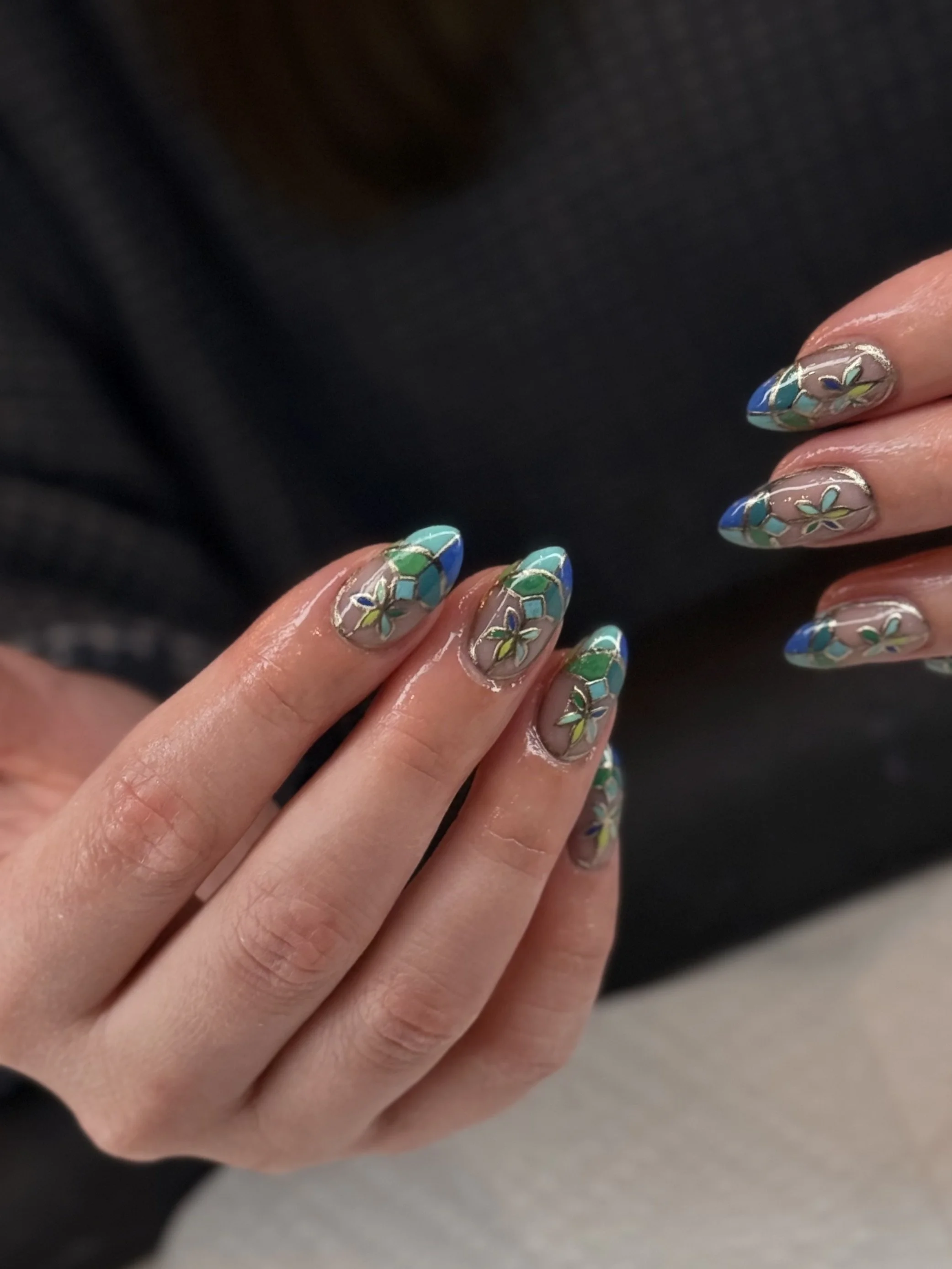 Close-up of a person's hand with manicured nails painted with blue and green floral nail art, against a dark background.