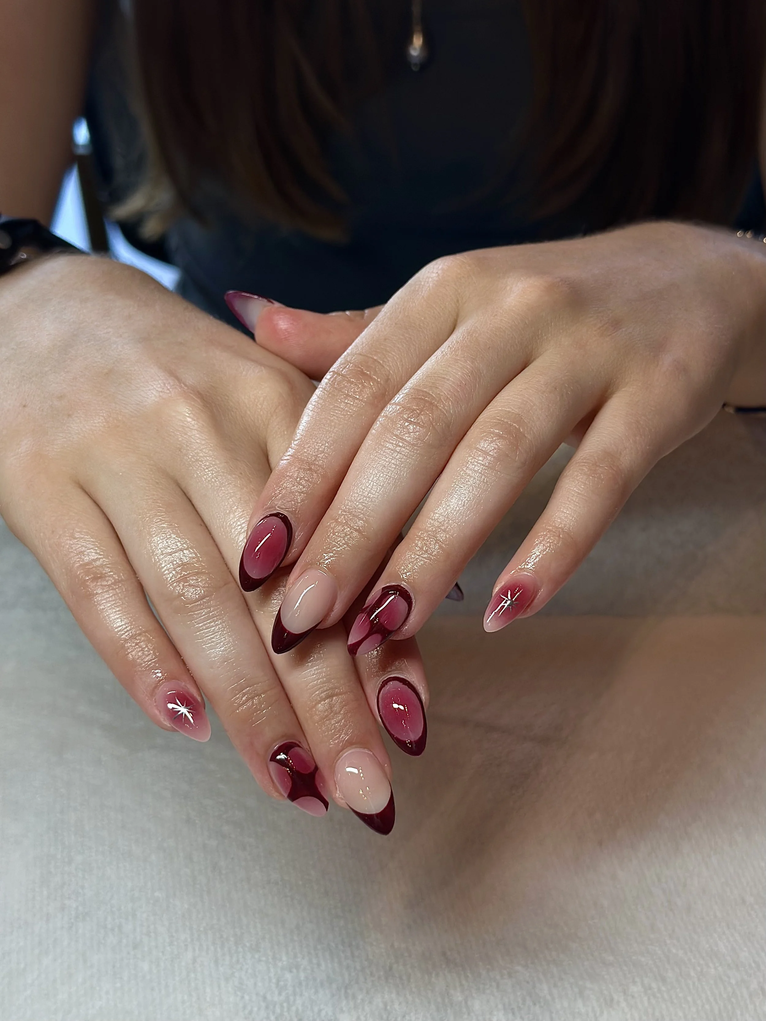 Close-up of a person's hands with manicured nails featuring a red and nude gel polish with white accents. The hands are resting on a light-colored surface, and the person's face and body are not visible.
