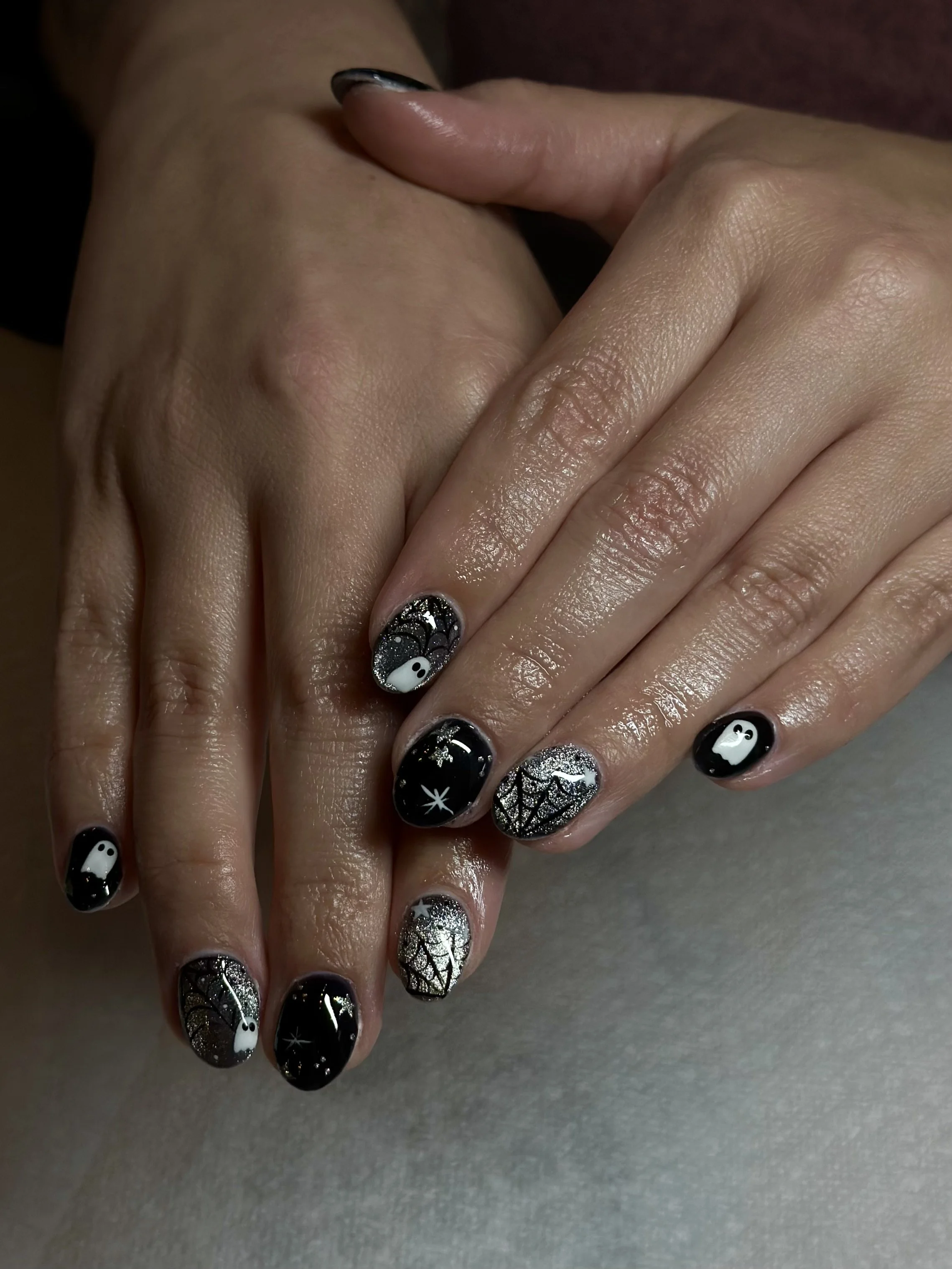 Close-up of hands with black and silver glittery Halloween-themed nail art, including bats, spiders, skulls, and ghost designs.