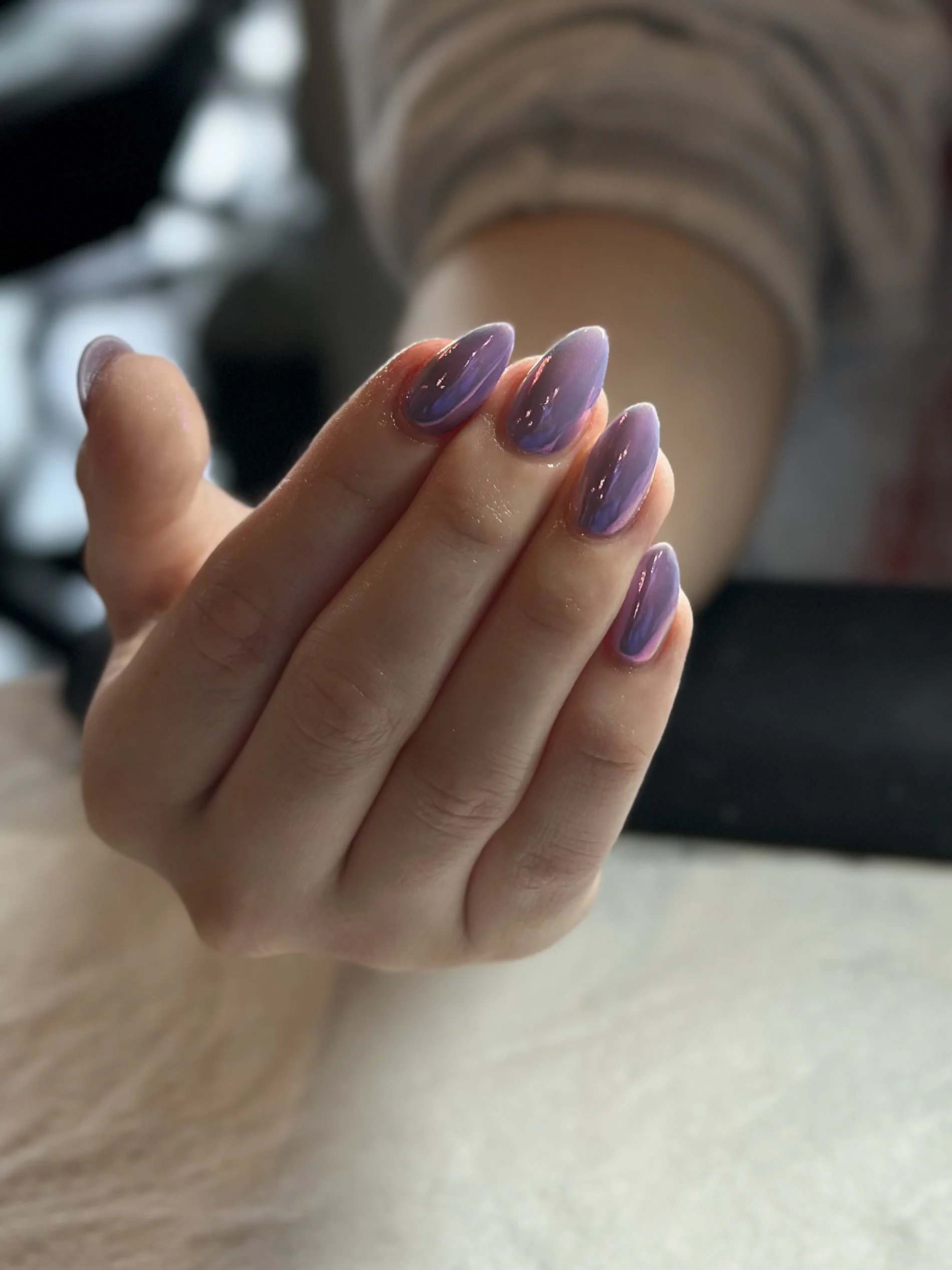 Close-up of a person's hand with manicured nails painted in purple, resting on a beige surface with blurred background of striped fabric.