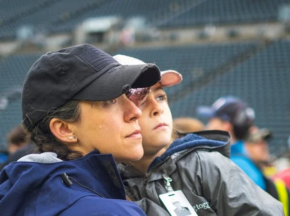 A woman and a young boy watching a sports event at a stadium, both wearing jackets and the woman wearing a black cap, with a blurred background of stadium seats and other spectators.