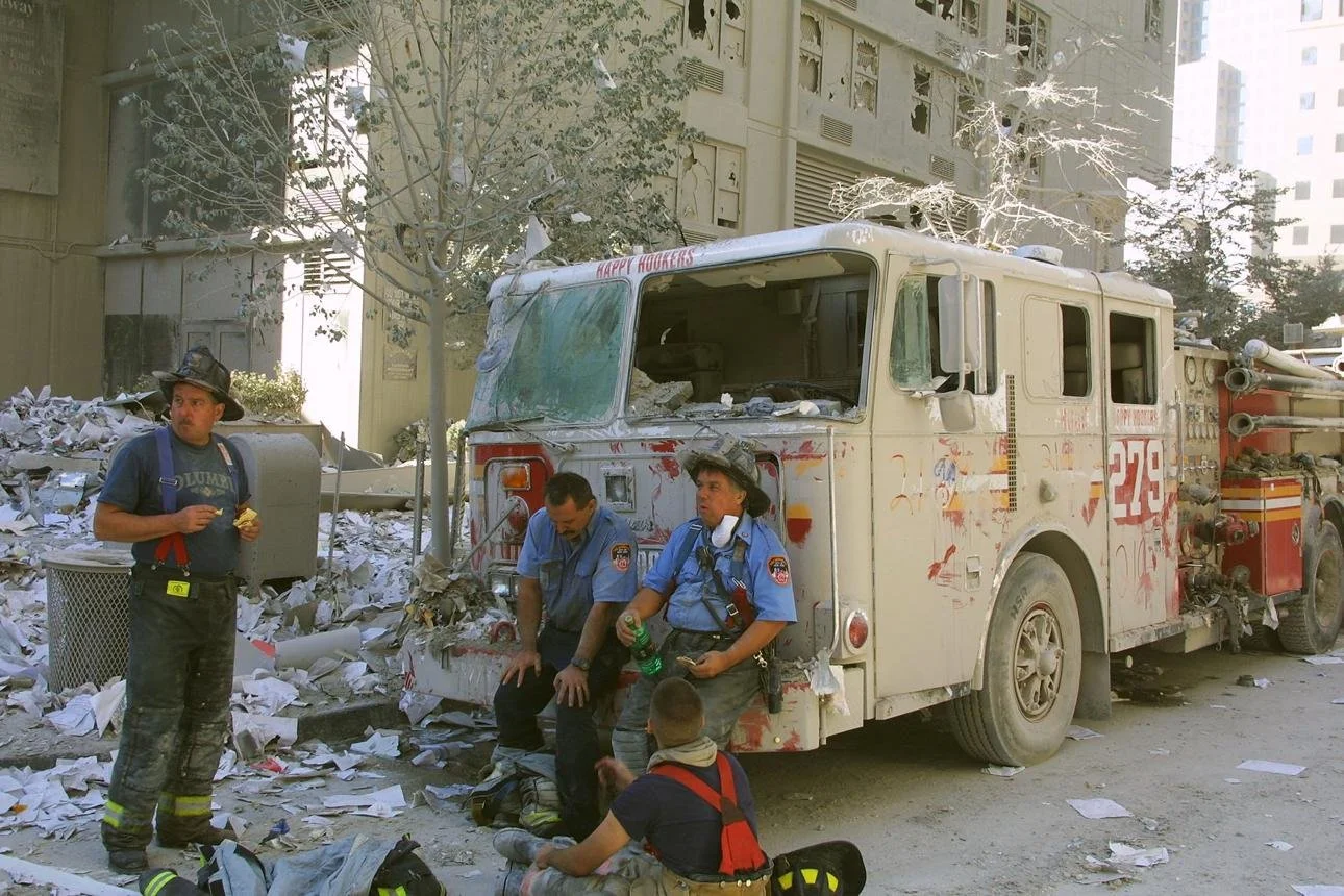 Four people, three in firefighter uniforms and one in civilian clothes, sit and stand near a damaged fire truck amid debris and trash in an urban setting.