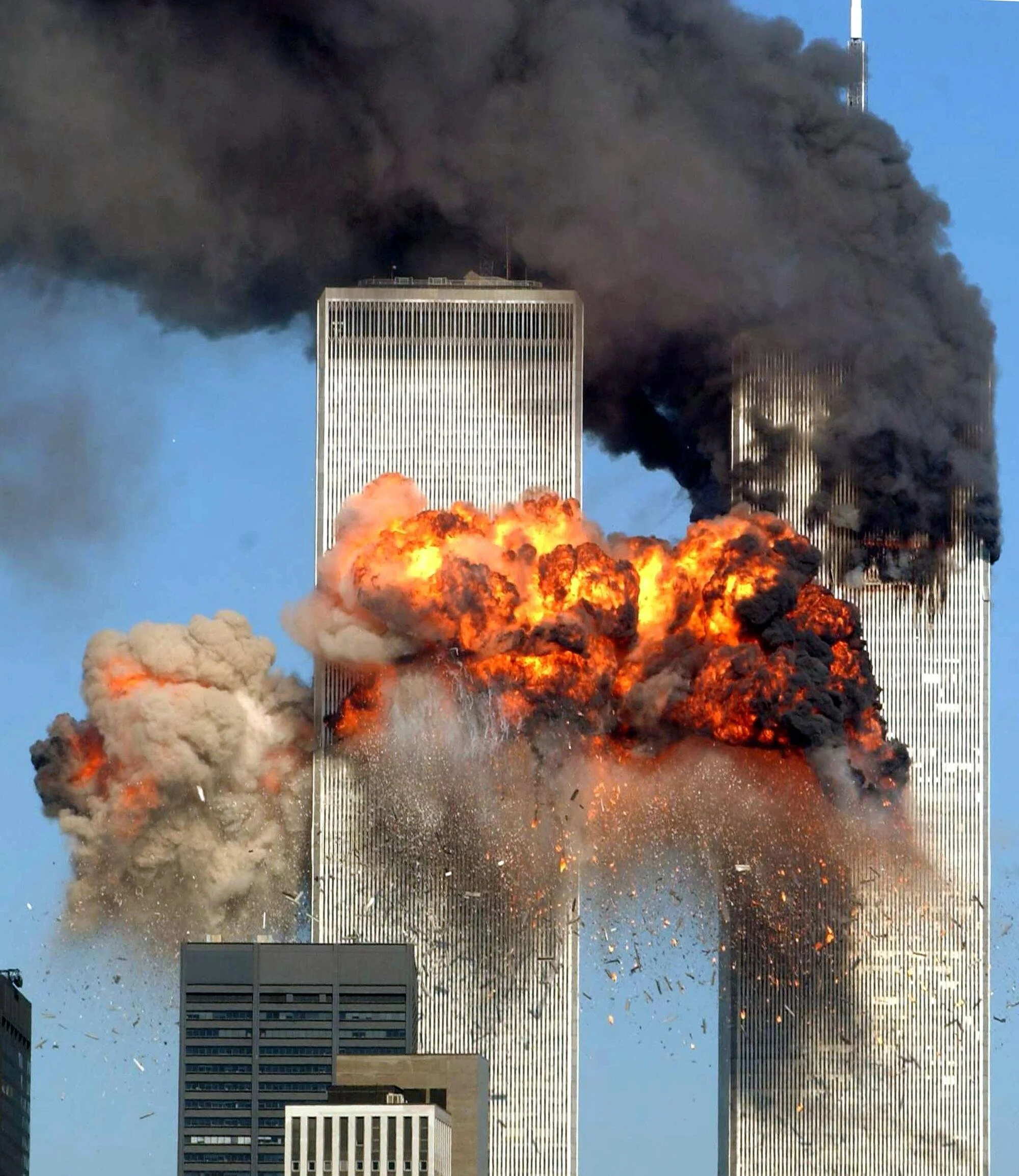 The World Trade Center Twin Towers engulfed in flames and thick black smoke during the September 11, 2001 terrorist attack, with debris falling and an adjacent building visible in the foreground.