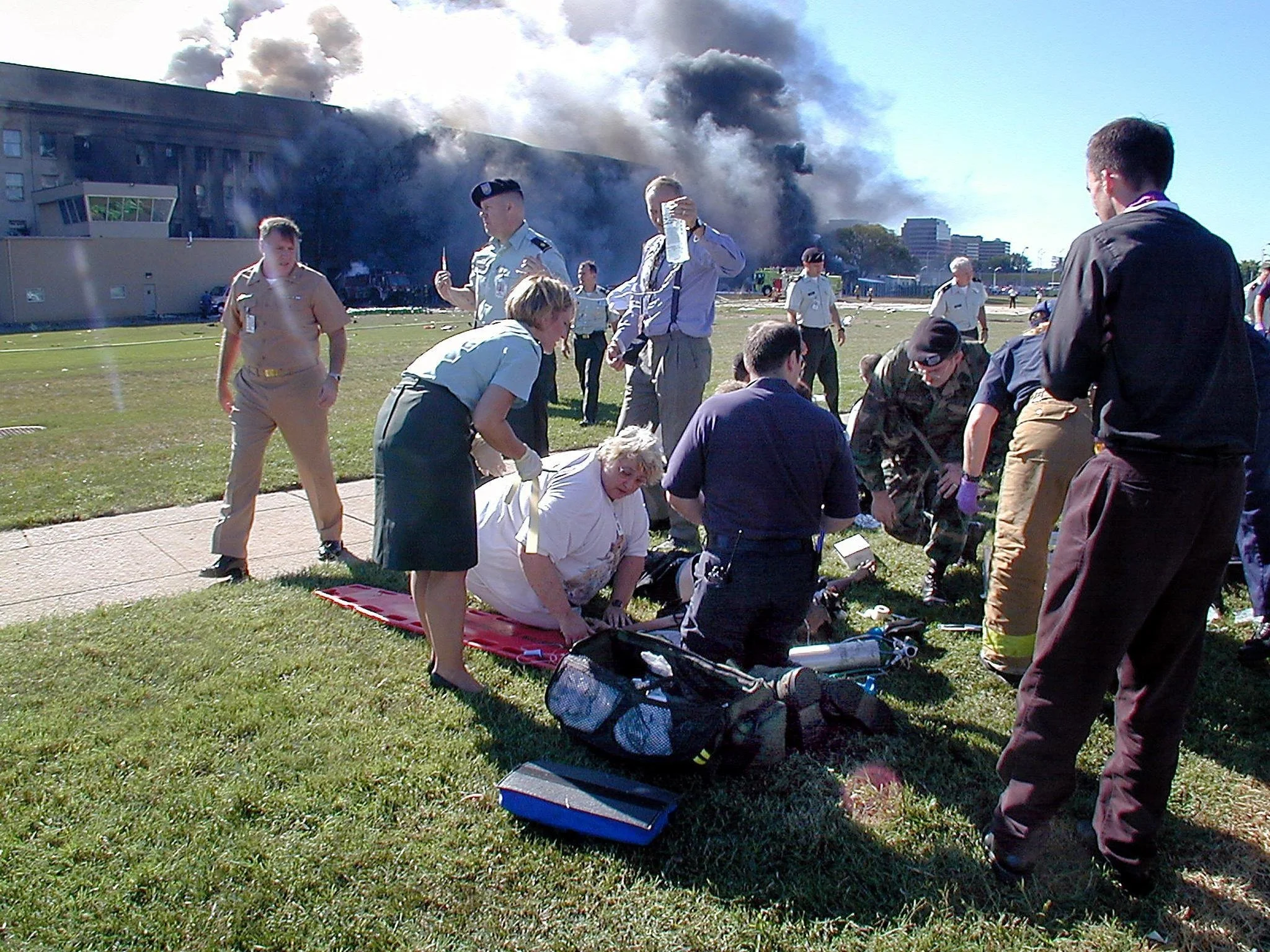 Scene of a fire emergency with smoke billowing from a building, surrounded by people including emergency responders and medical personnel attending to an injured individual on the grass.